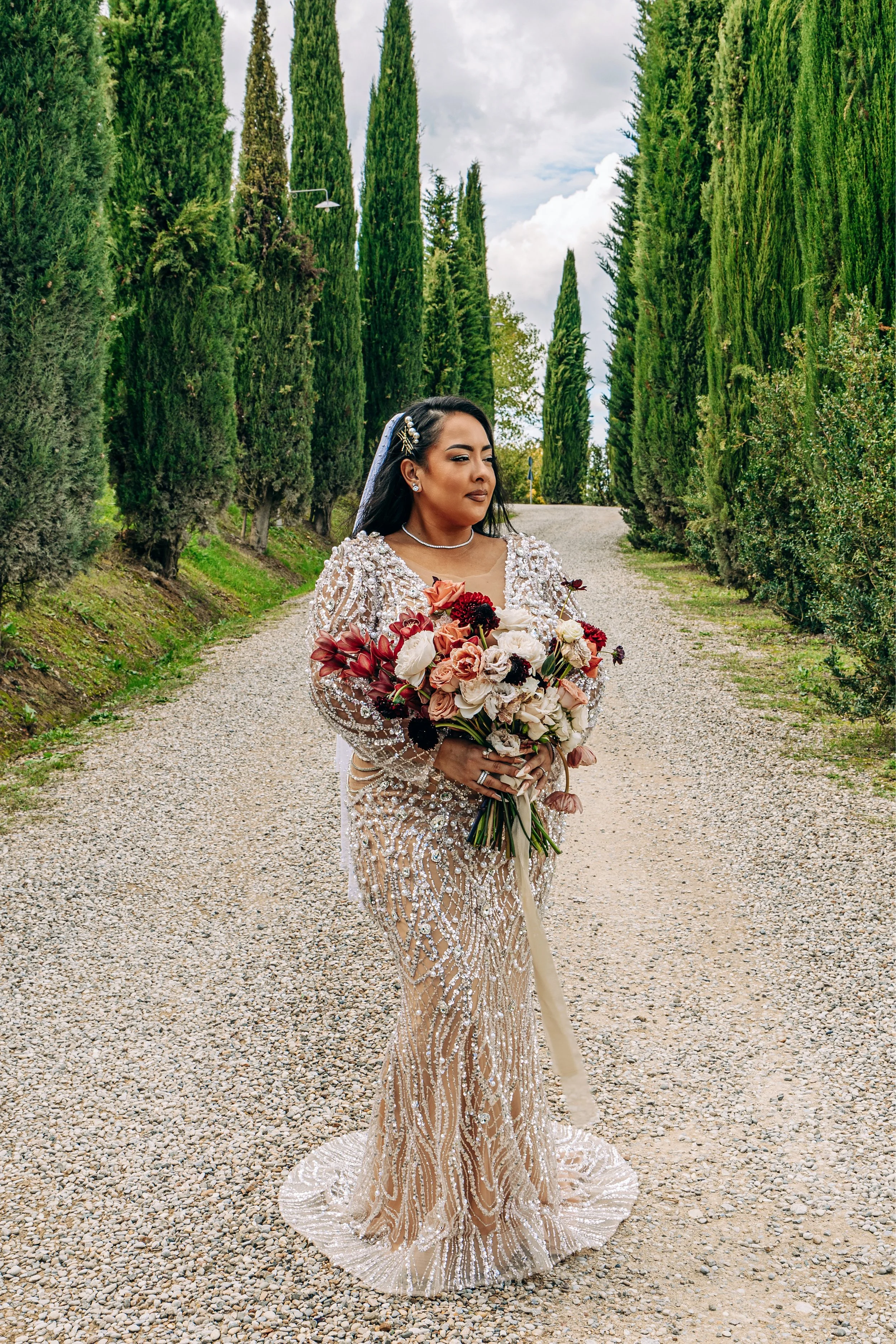 A woman in an ornate, silver-beaded engagement dress holding a large bouquet of flowers, standing on a gravel path surrounded by tall green cypress trees under a partly cloudy sky.