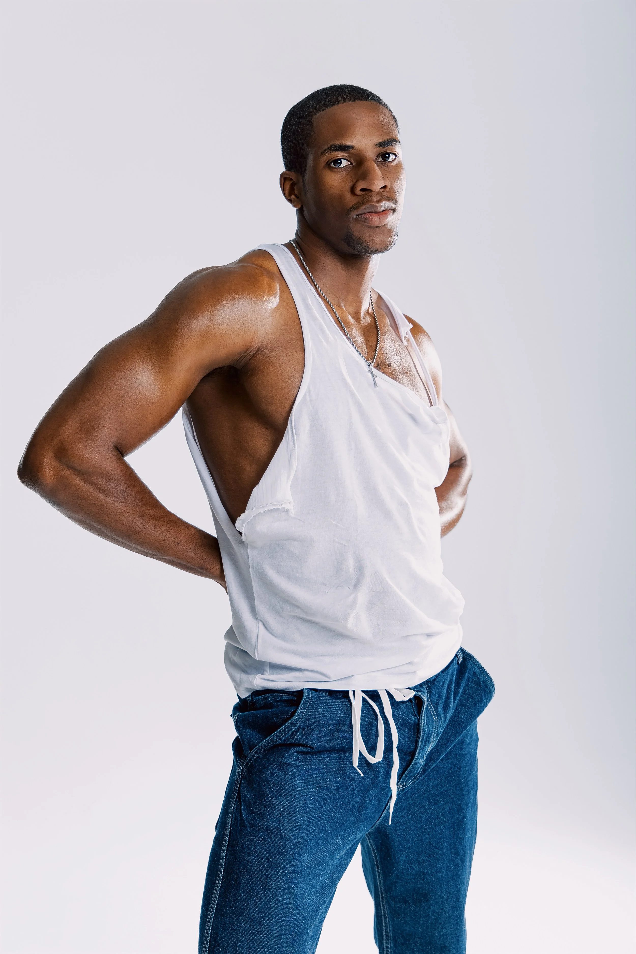 A young man with short black hair, wearing a sleeveless white tank top and blue jeans, standing against a plain light gray background.