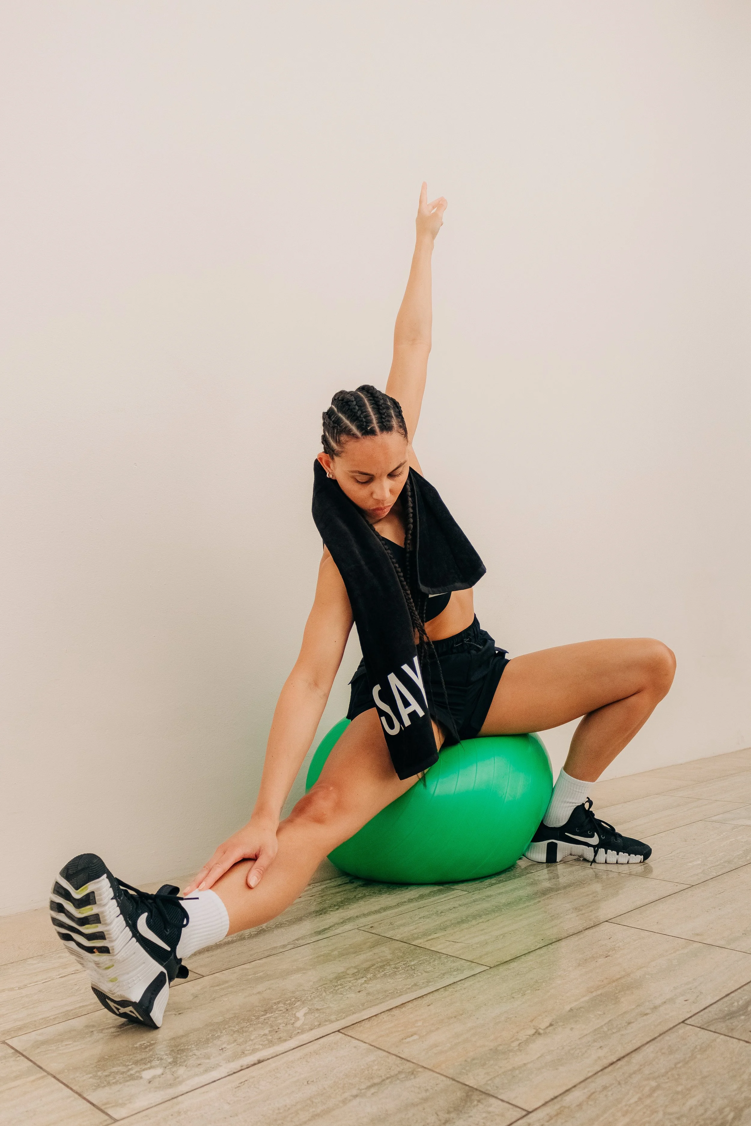 A young woman exercising indoors, sitting on a green exercise ball, stretching her leg with one arm and raising the other arm upwards, against a plain white wall.