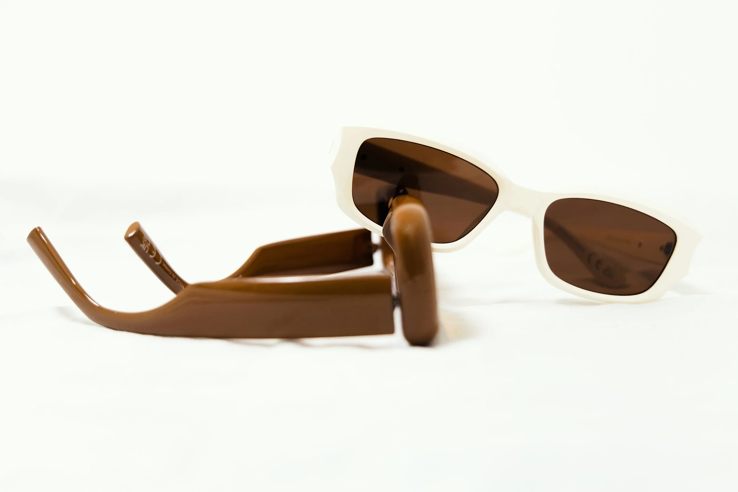 Pair of sunglasses with a white frame and brown lenses, resting on a brown case, against a white background.
