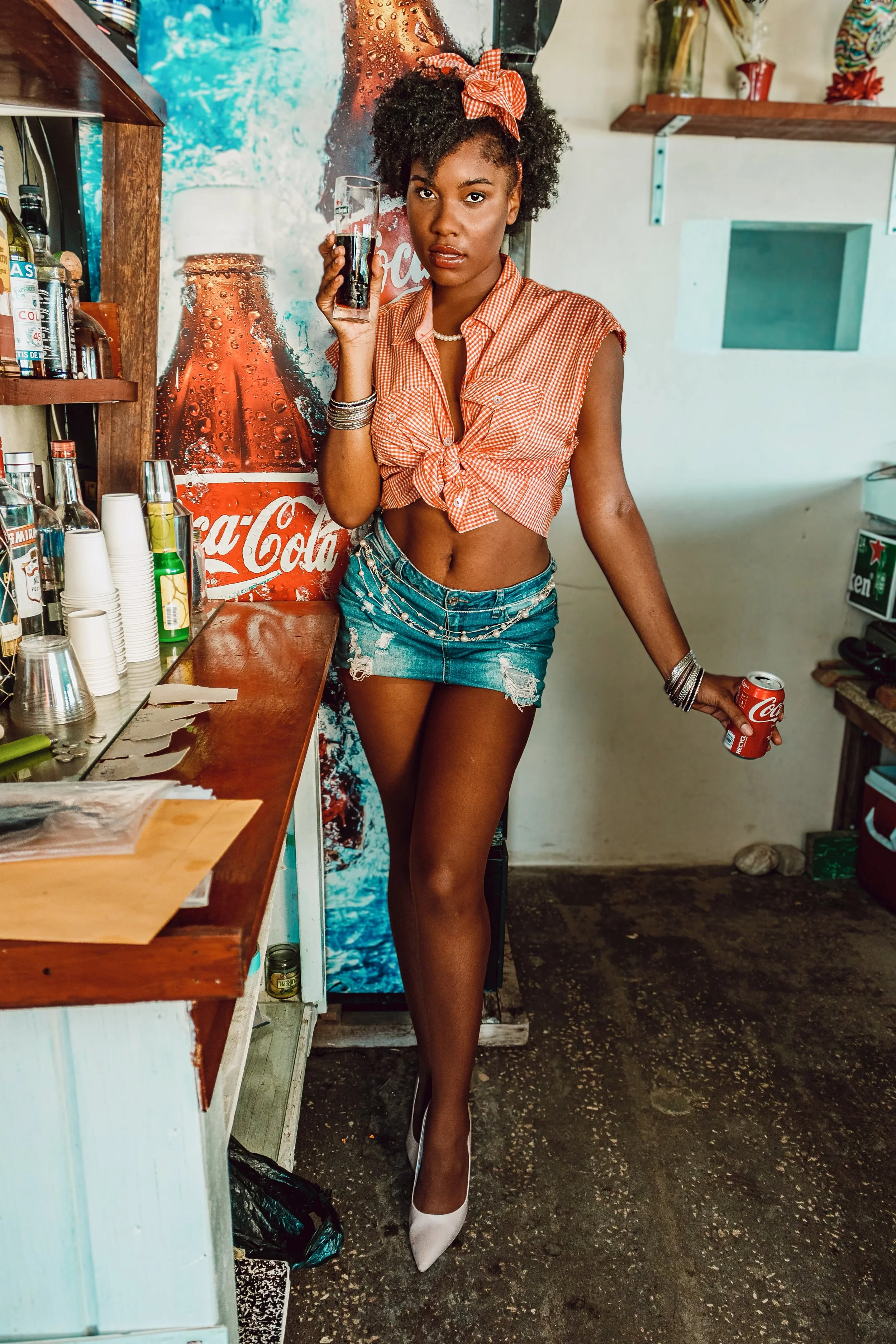 A woman with curly hair, wearing a red gingham sleeveless shirt tied at the waist, distressed denim shorts, white high heels, pearl necklace, and bracelets. She holds a glass of Coca-Cola in one hand and a soda can in the other, standing next to a Co
