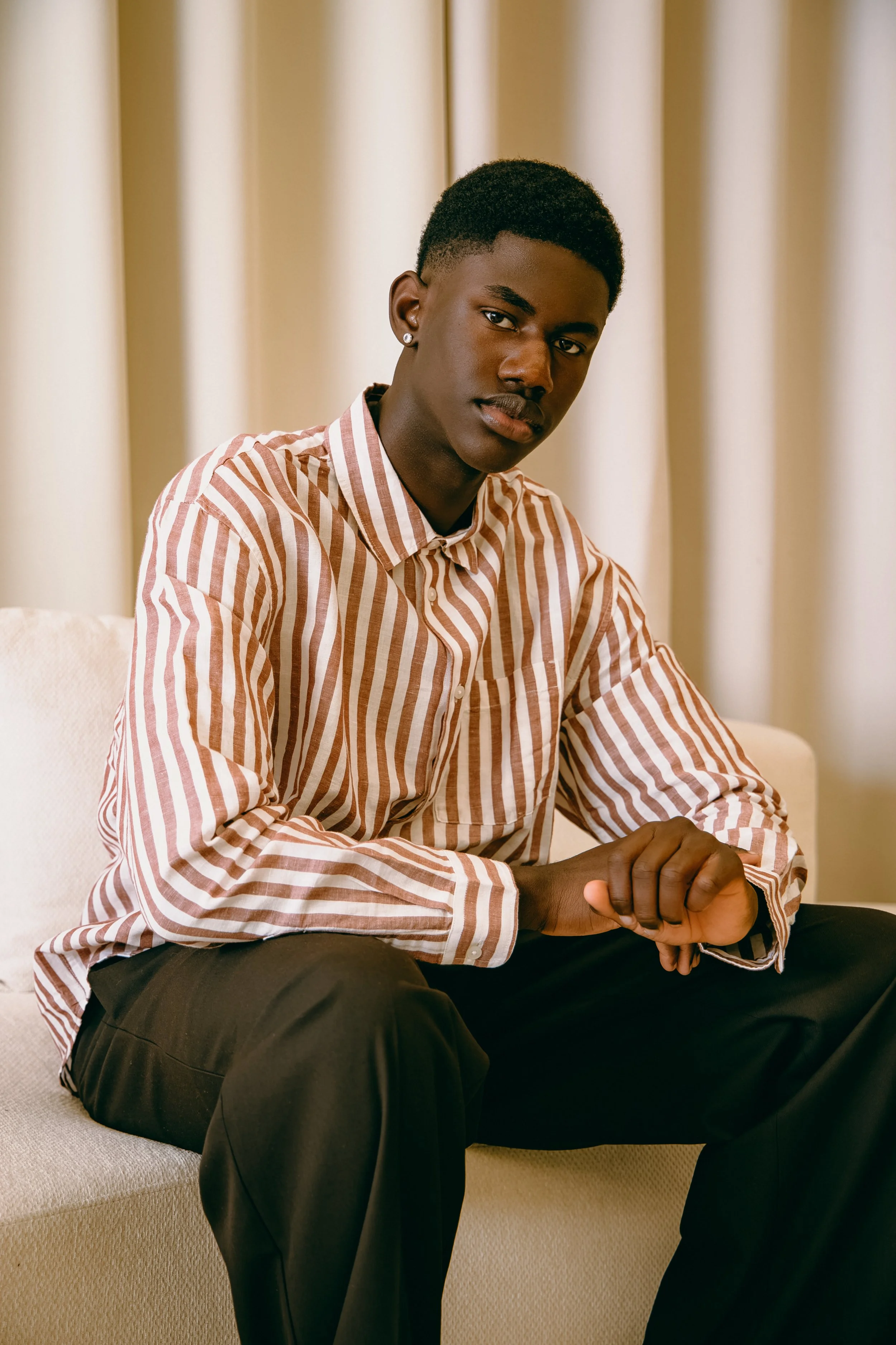 A young Black man sitting on a beige sofa in front of cream-colored curtains, wearing a striped button-up shirt and black pants, with a neutral expression and his hands clasped.