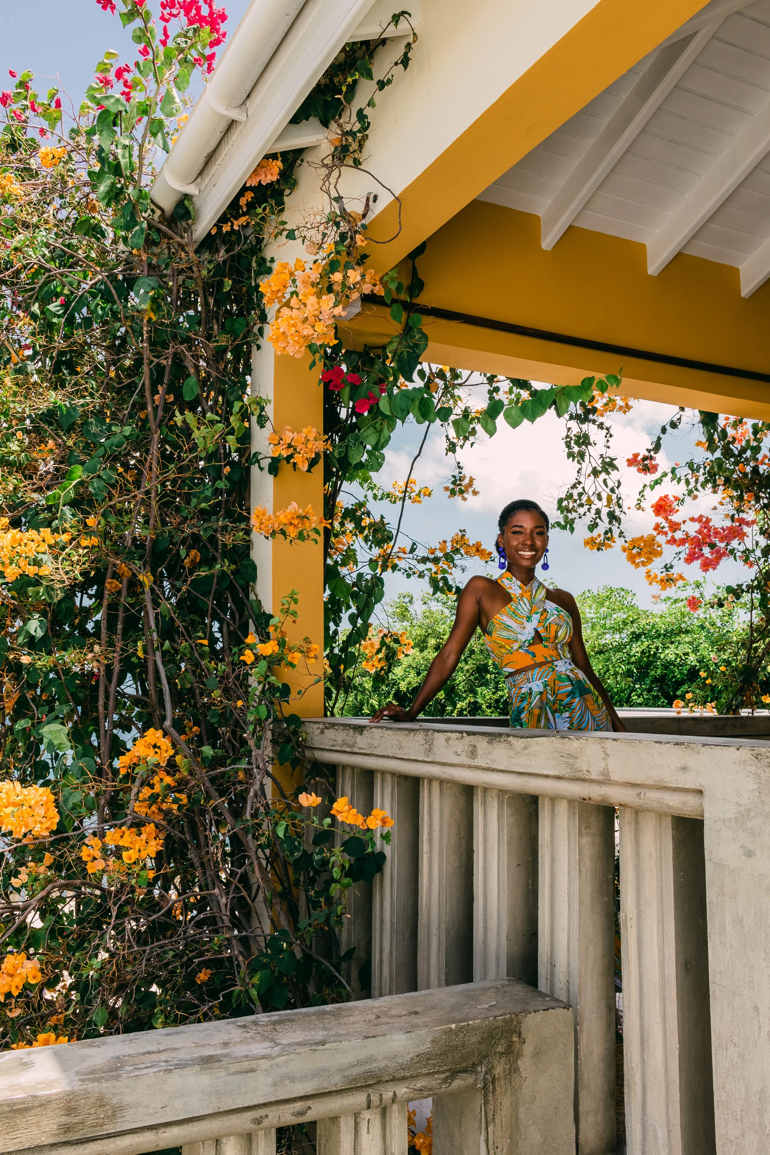A woman in a colorful, patterned dress standing on a balcony, surrounded by flowering plants with orange, pink, and green foliage, under a yellow and white pergola with a cloudy sky above.