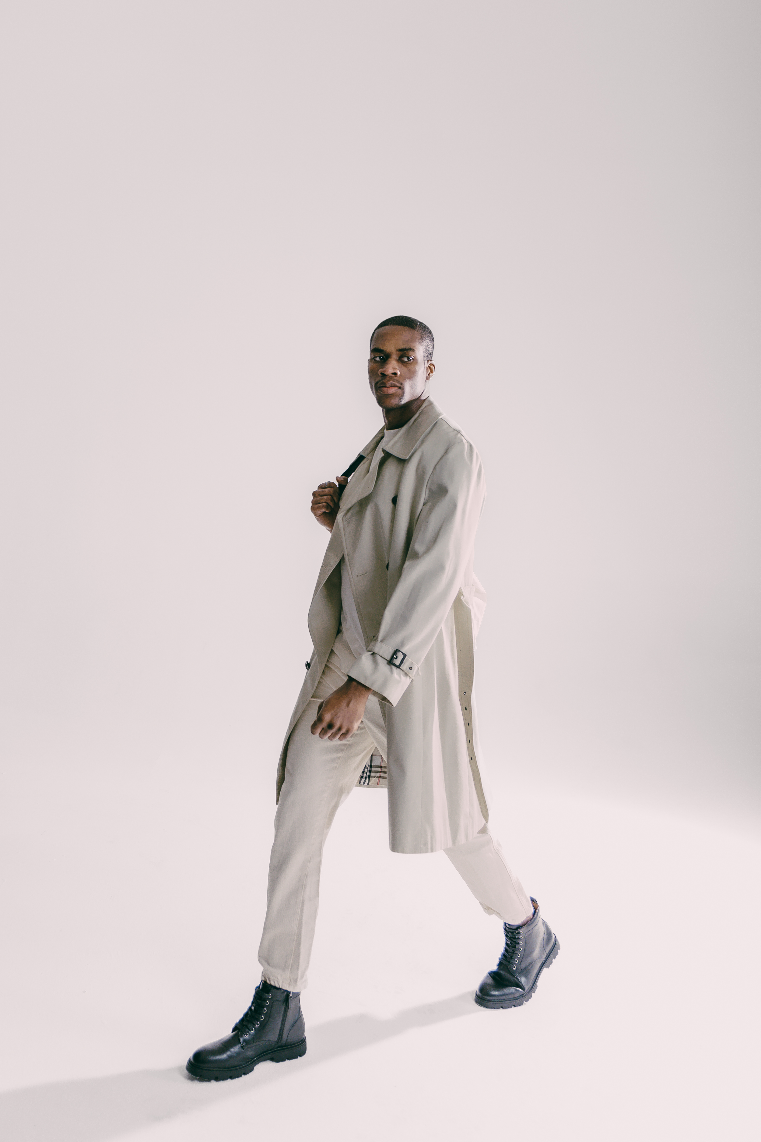 A young man walking in a studio with a plain white background, wearing a beige trench coat, beige pants, black combat boots, and carrying a black bag over his shoulder.