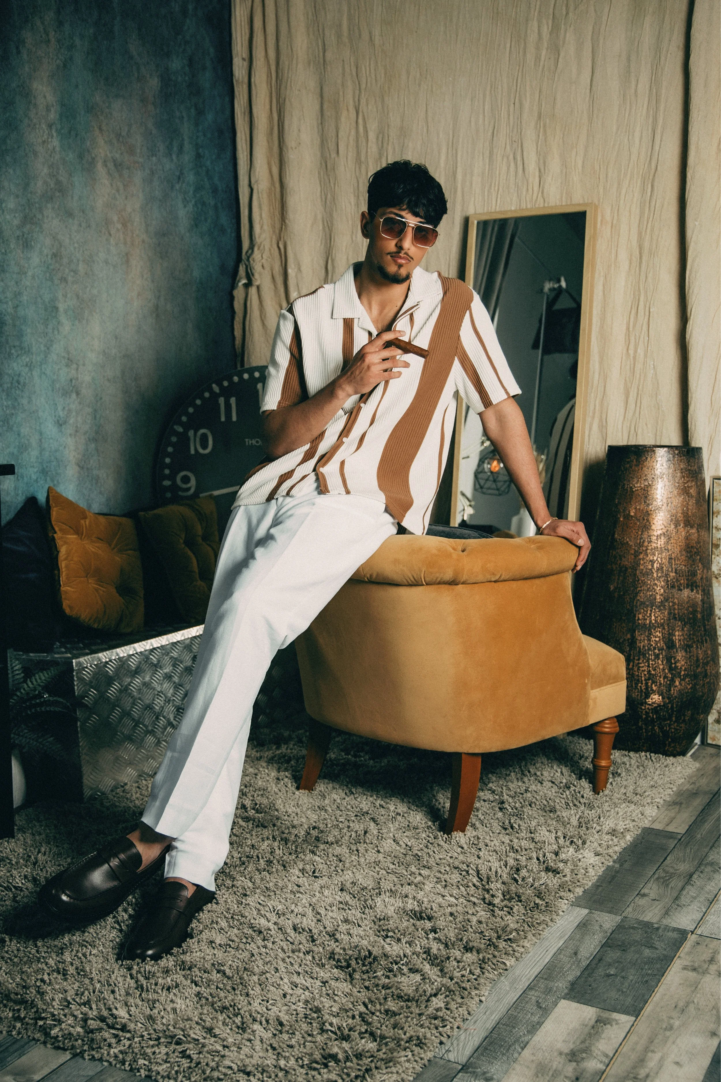 A young man with dark hair, sunglasses, and facial hair, dressed in a striped shirt and white pants, stands in a vintage-style living room, smoking a cigar while leaning on a mustard-colored armchair.