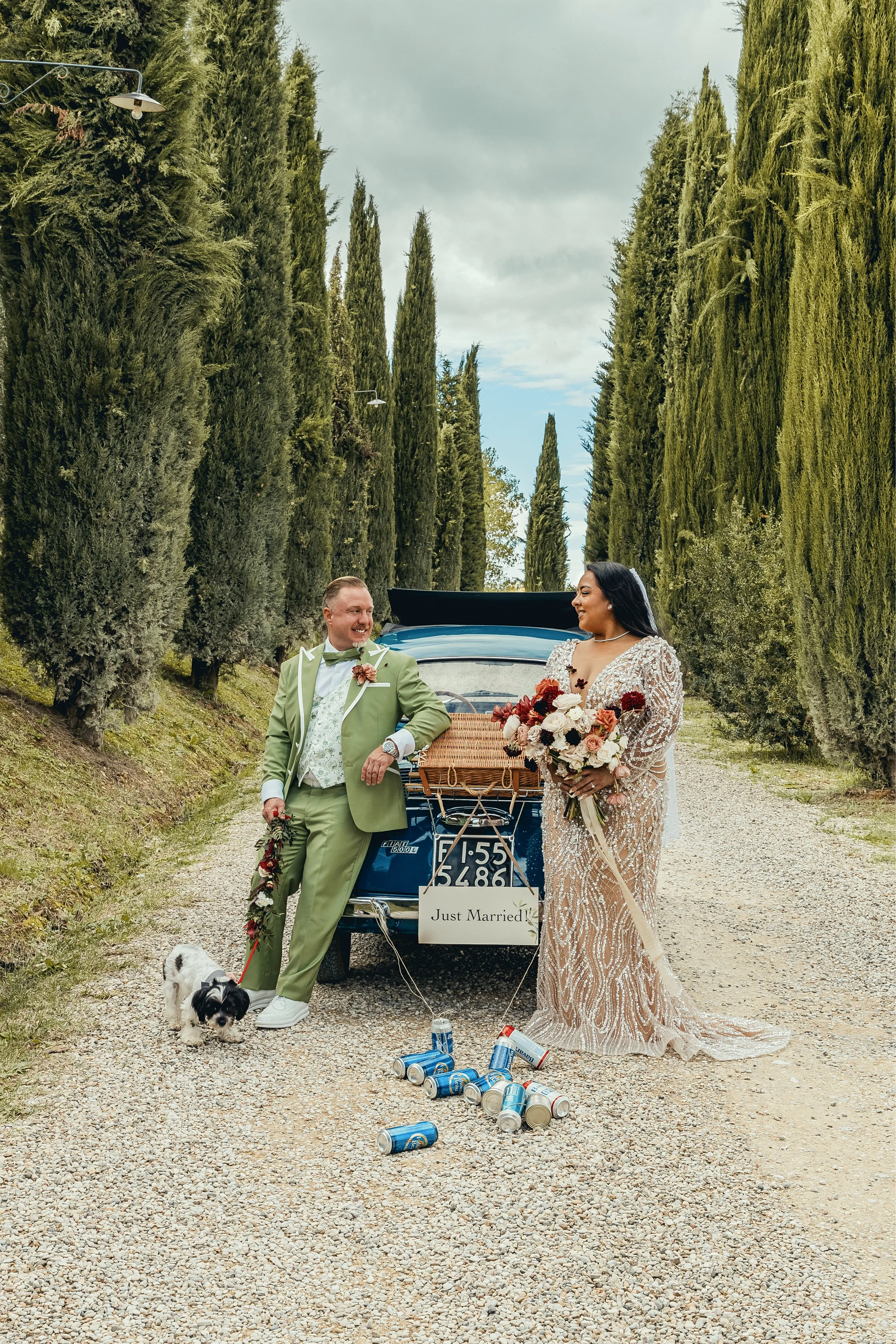 A newly married couple smiling at each other outdoors, standing in front of a vintage blue car with a 