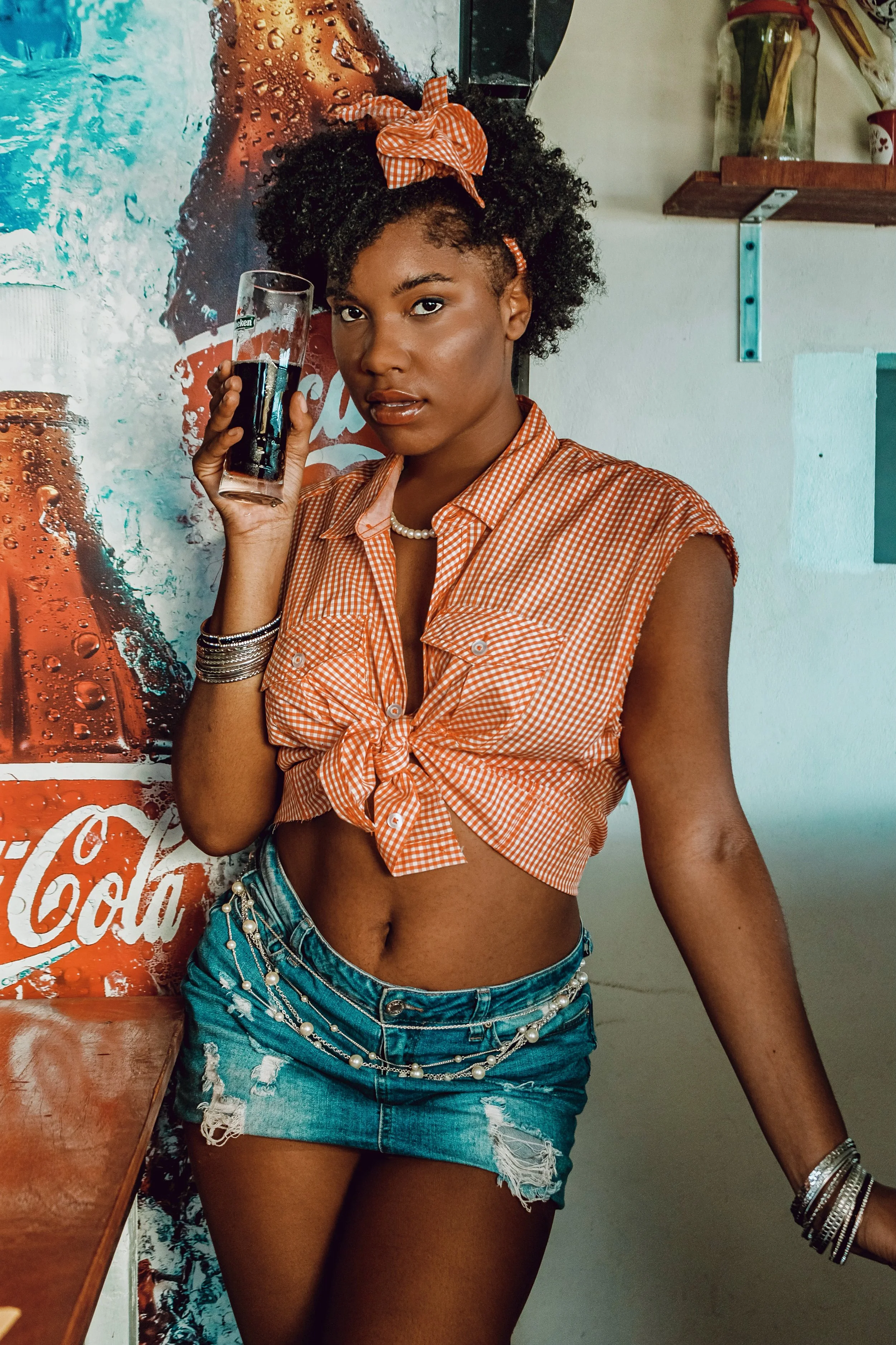 A young woman with curly hair wearing a orange checkered sleeveless shirt tied at the waist, denim shorts with pearl chains, holding a drink, and standing in a retro decor setting.