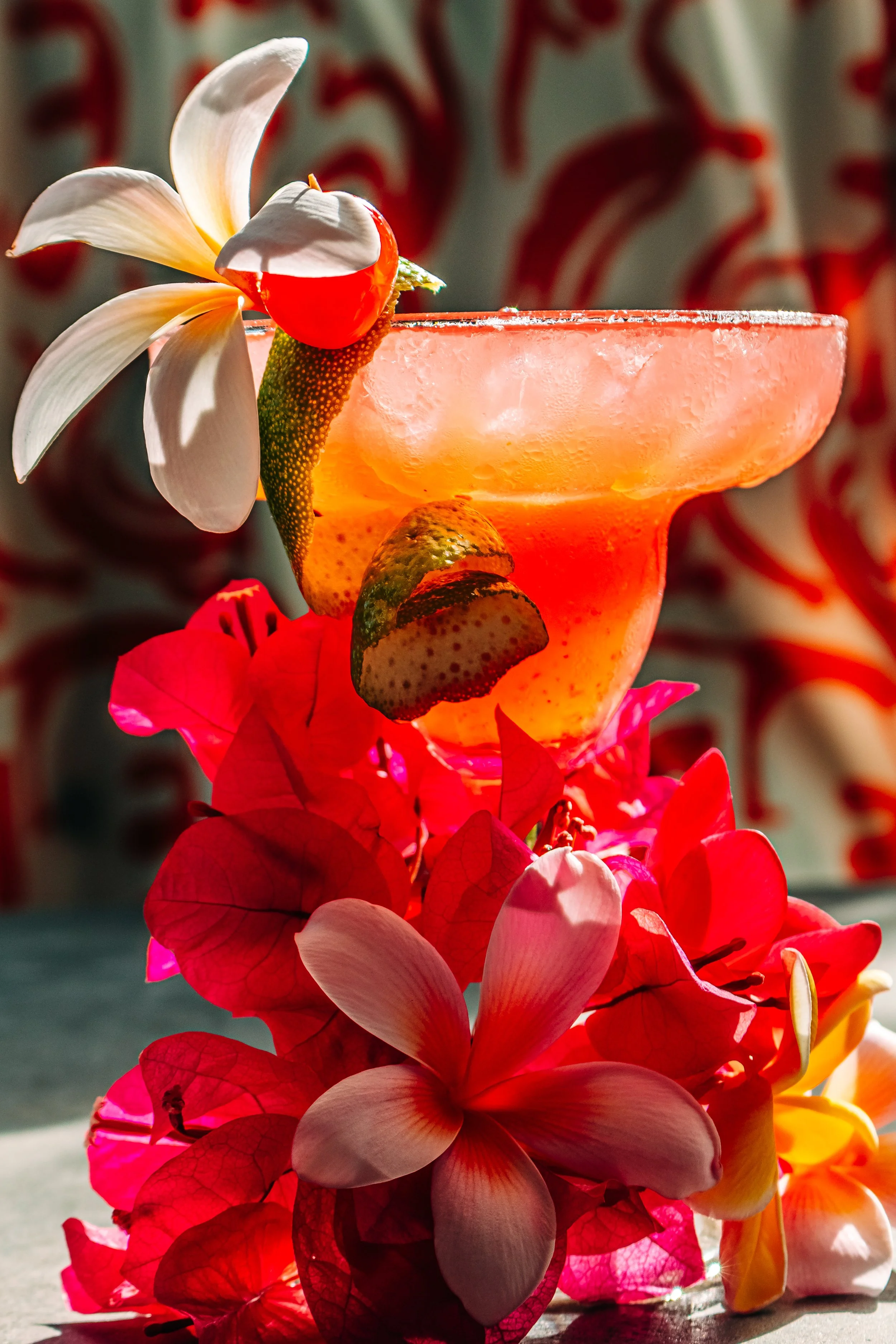 A colorful cocktail in a margarita glass decorated with a red cherry, a white flower, a slice of lime, and pink bougainvillea flowers at the base, with a red and white patterned curtain in the background.