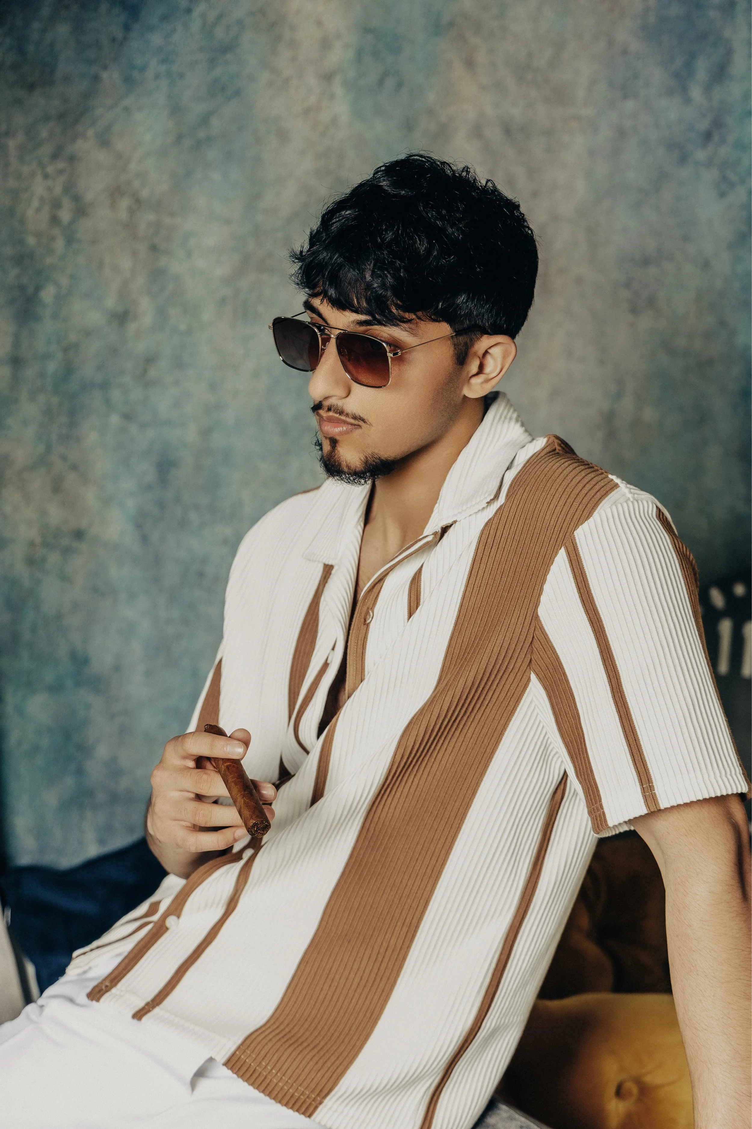 A young man with dark hair, sunglasses, and a small beard, wearing a striped short-sleeved shirt in white and brown, holding a cigar in his right hand, sitting against a textured background.