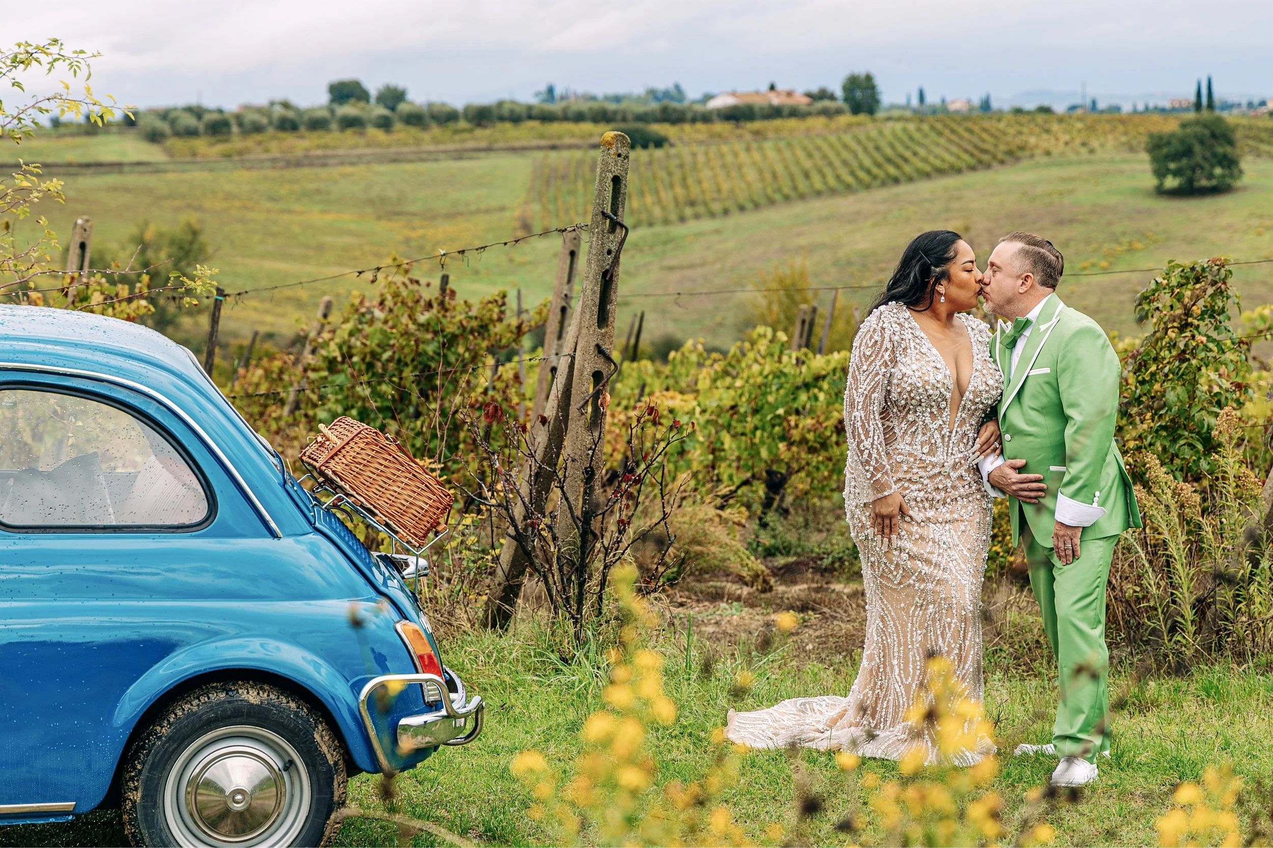 A couple is kissing in a vineyard with a vintage blue car parked nearby. The woman is wearing an embellished gown, and the man is in a bright green suit. The scene is set outdoors with rolling hills, grapevines, and a cloudy sky in the background.