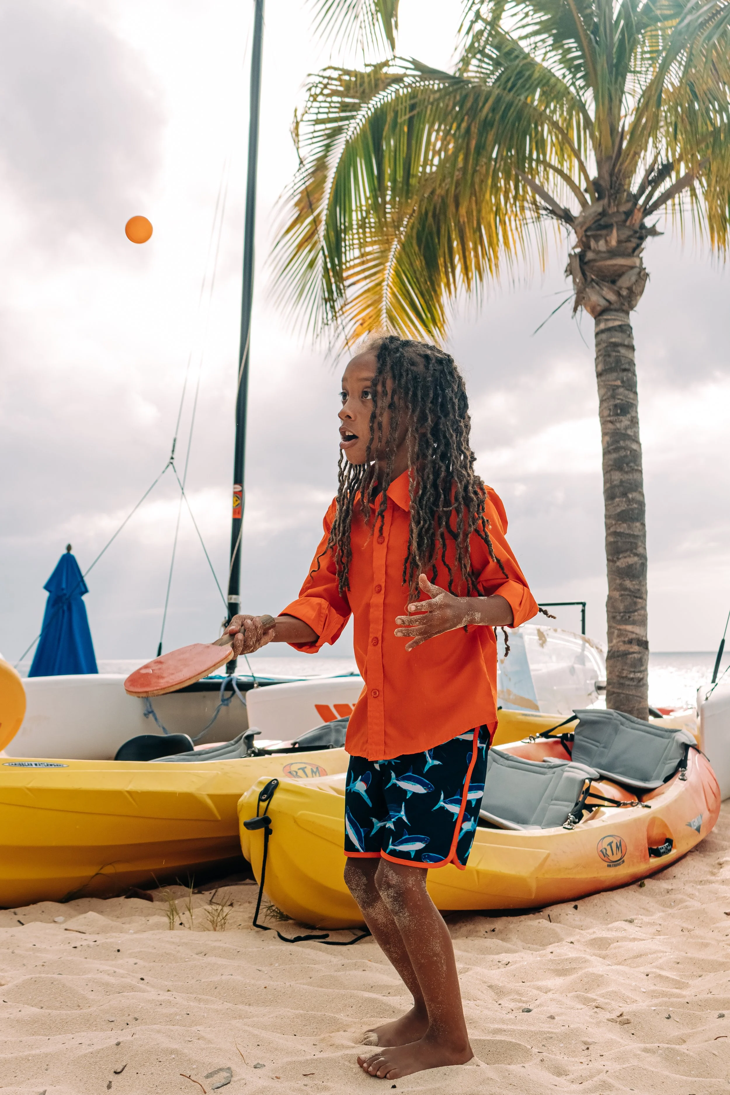 A young boy with long, curly hair wearing an orange shirt and shorts with fish patterns, standing on a sandy beach, holding a paddle, with yellow kayaks and palm trees in the background.