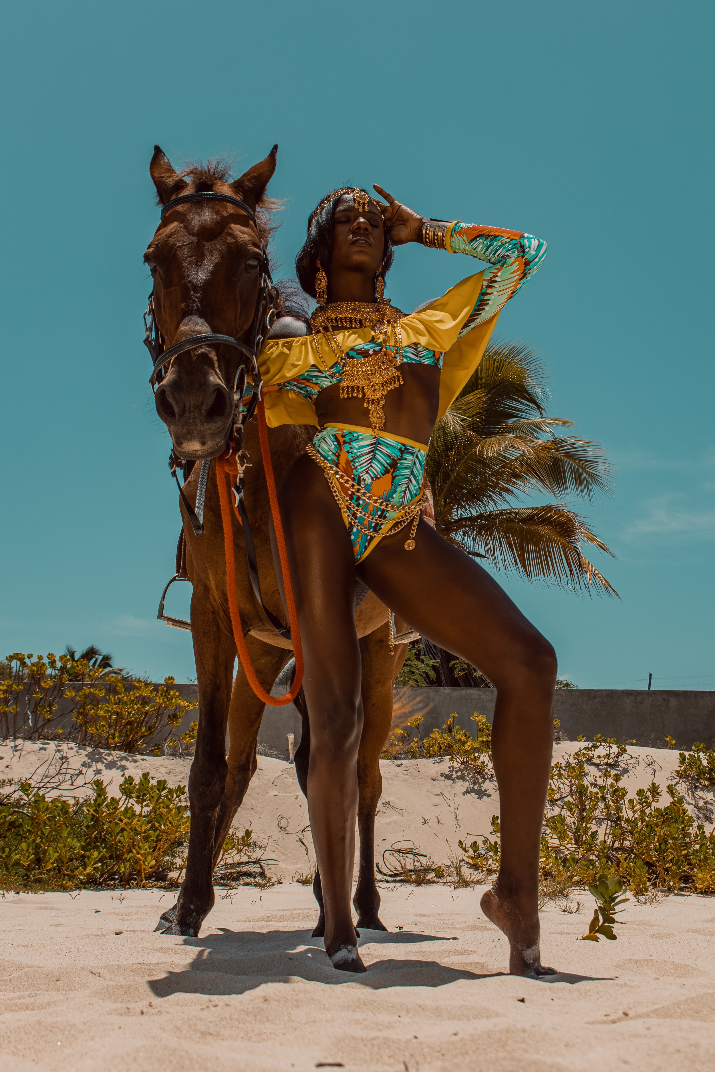 A woman in colorful, tropical-themed clothing and gold jewelry standing beside a horse on a sandy beach with palm trees and a clear blue sky in the background.
