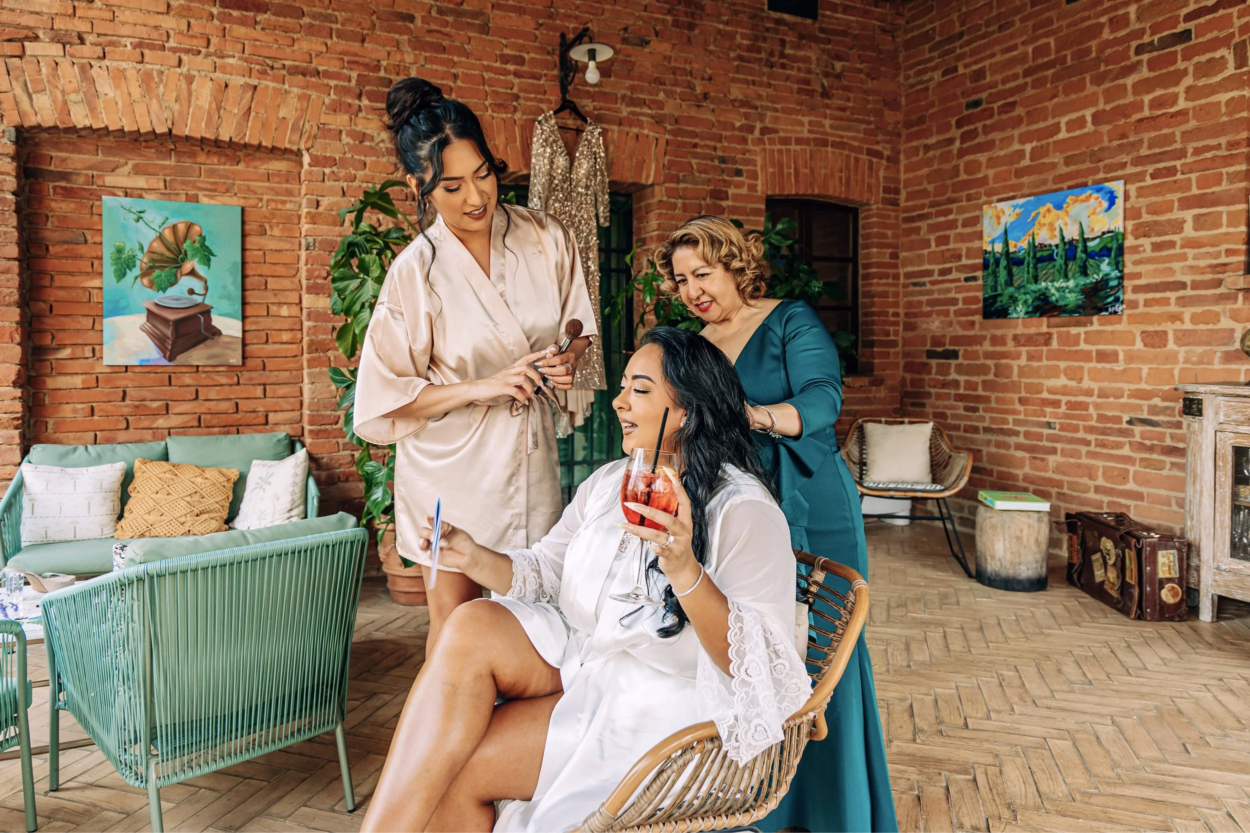 Four women in a cozy brick-walled room, one sitting with a drink, while others prepare her makeup.