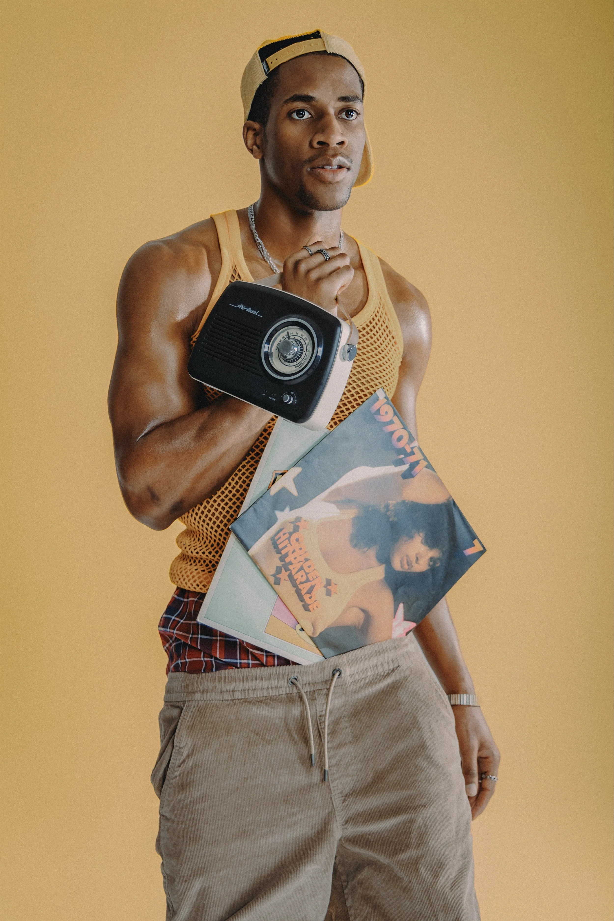 A young man in a yellow tank top and beige shorts holding a vinyl record and a vintage portable radio, standing against a plain beige background.
