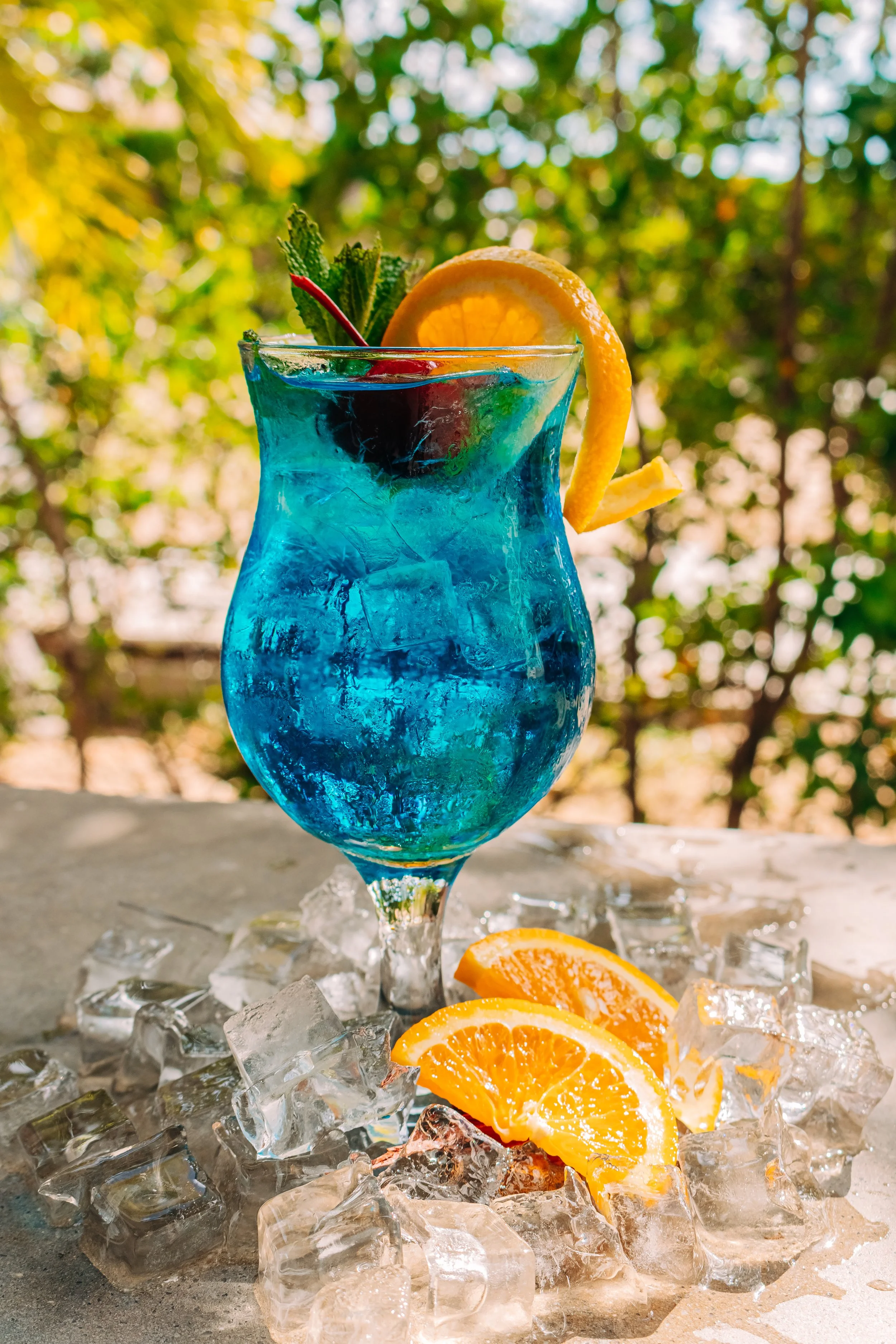 A blue cocktail with ice cubes, garnished with a lemon slice, a cherry, and a mint sprig, sitting on ice and sliced oranges on a table outdoors with a blurred green leafy background.