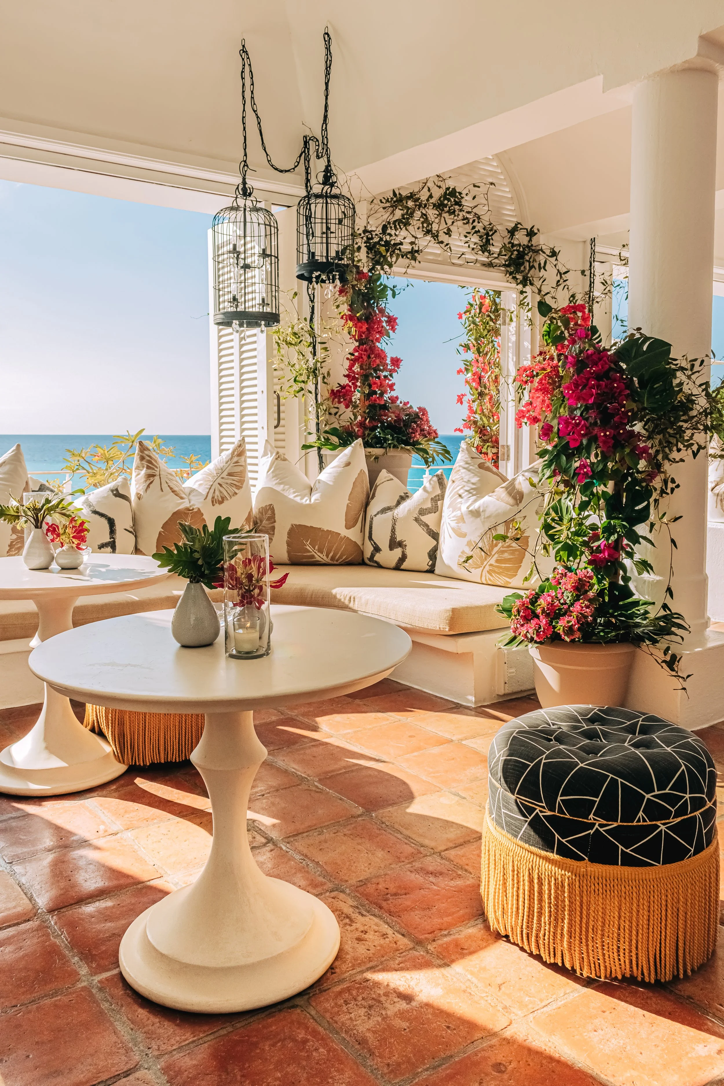 A sunlit terrace with terra cotta tiled floor, featuring a seating area with pillows, a small round table with vases and flowers, lush greenery with pink flowers, and a view of the ocean through open white shutters.