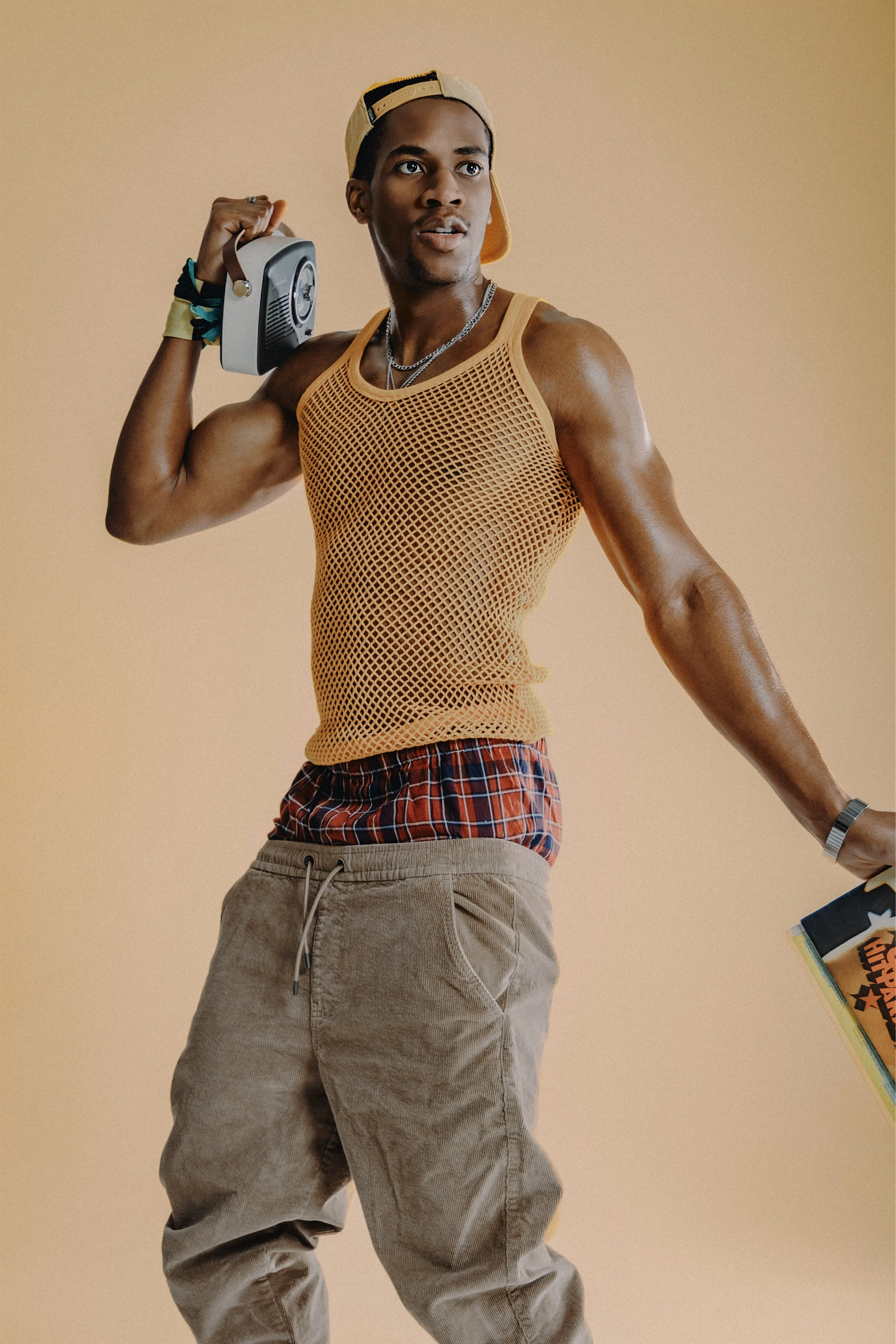 A young man in a yellow mesh tank top, plaid underwear, beige pants, and a backwards cap, holding a boombox on his shoulder and a book in his other hand, standing against a beige background.