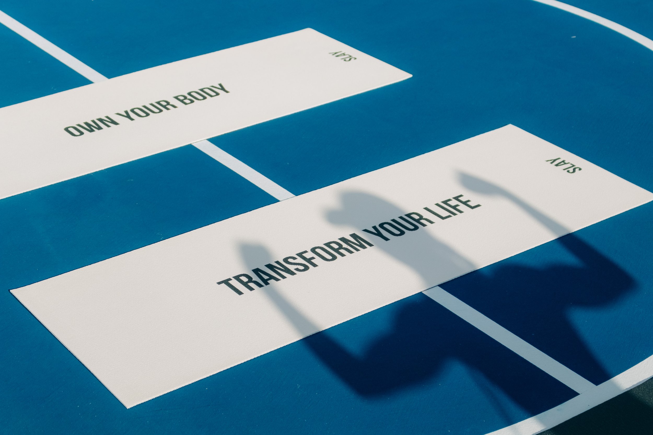 A blue life-size game board with white spaces labeled "Own Your Body" and "Transform Your Life" in black text.
