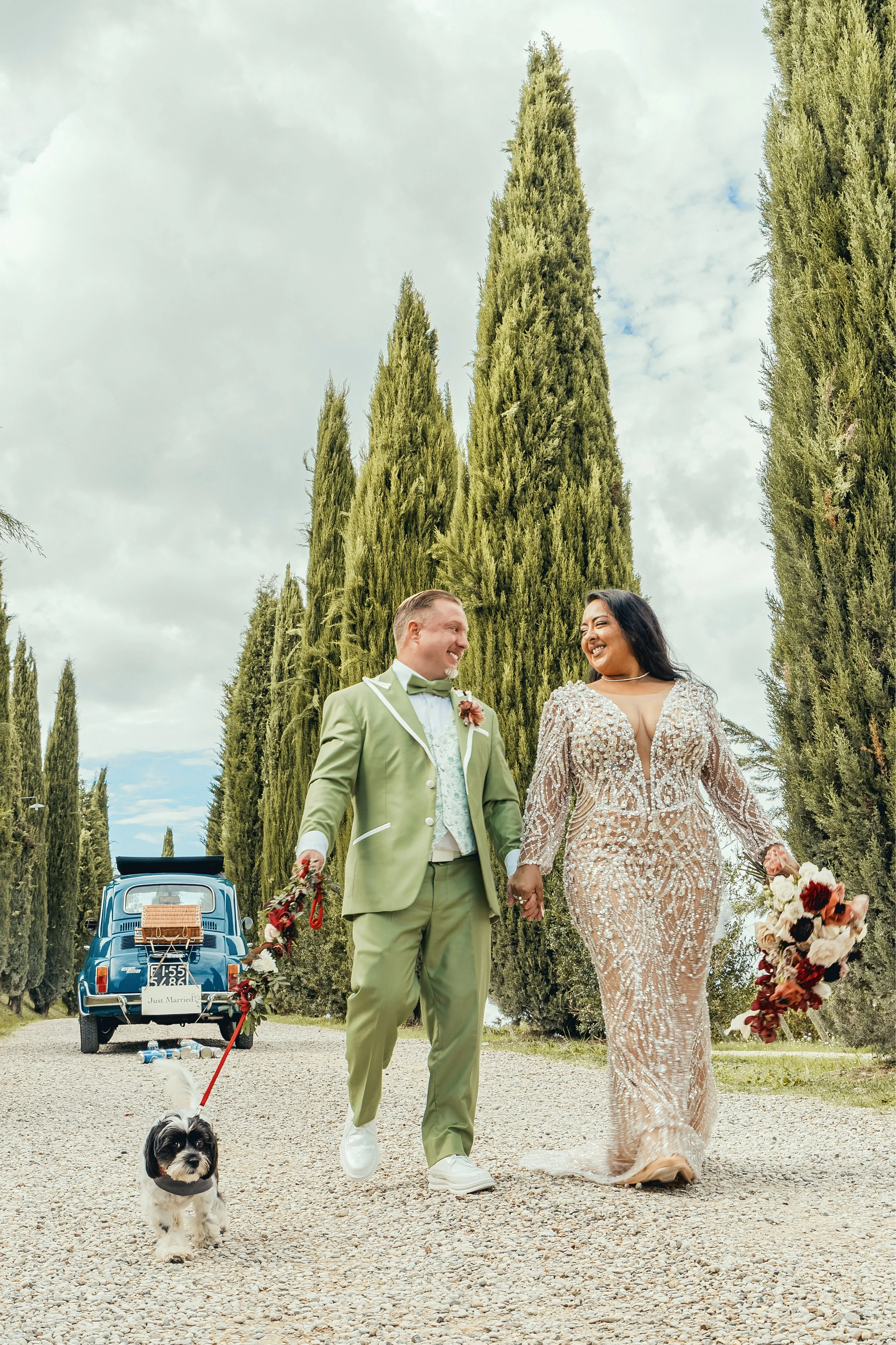 A newly married couple walking hand in hand in an outdoor setting with tall cypress trees. The groom is wearing a light green suit with a bow tie, and the bride is wearing a shimmering, intricate, long dress with a bouquet of flowers. A small dog on 