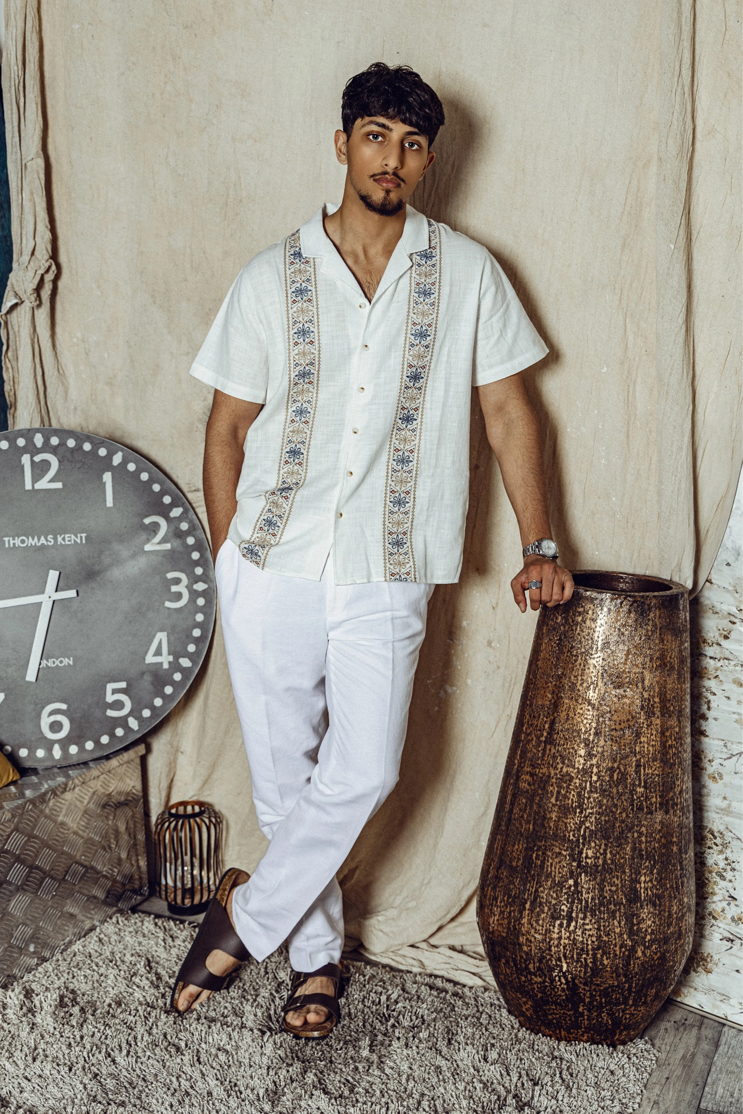 A young man standing indoors against a beige backdrop, wearing a white short-sleeve shirt with embroidered details, white pants, and sandals, resting his left hand on a large bronze-colored vase, with a large wall clock nearby.
