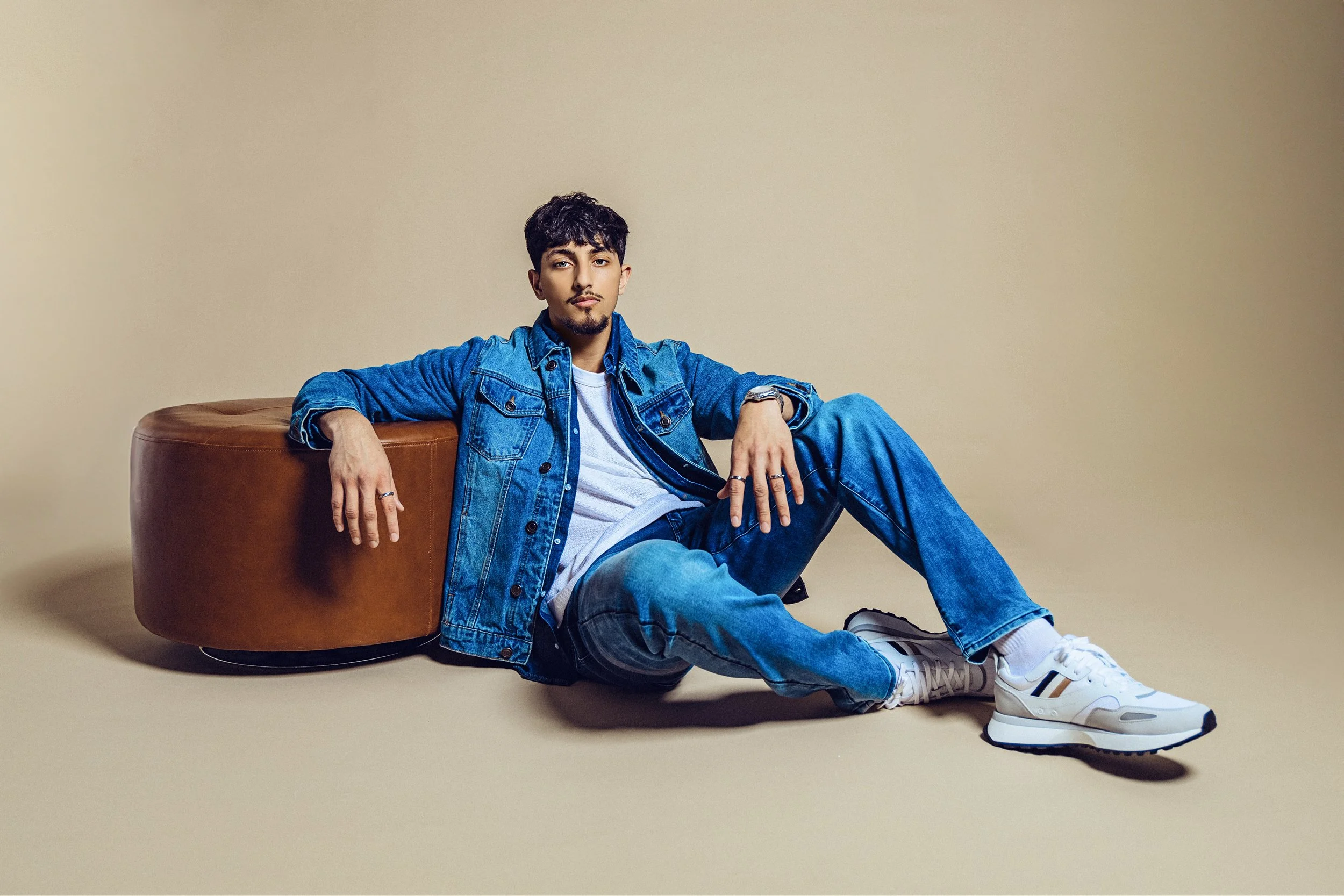A young man wearing a denim jacket and jeans, white sneakers, and a watch, sitting on the floor with his back against a brown leather ottoman, against a beige background.