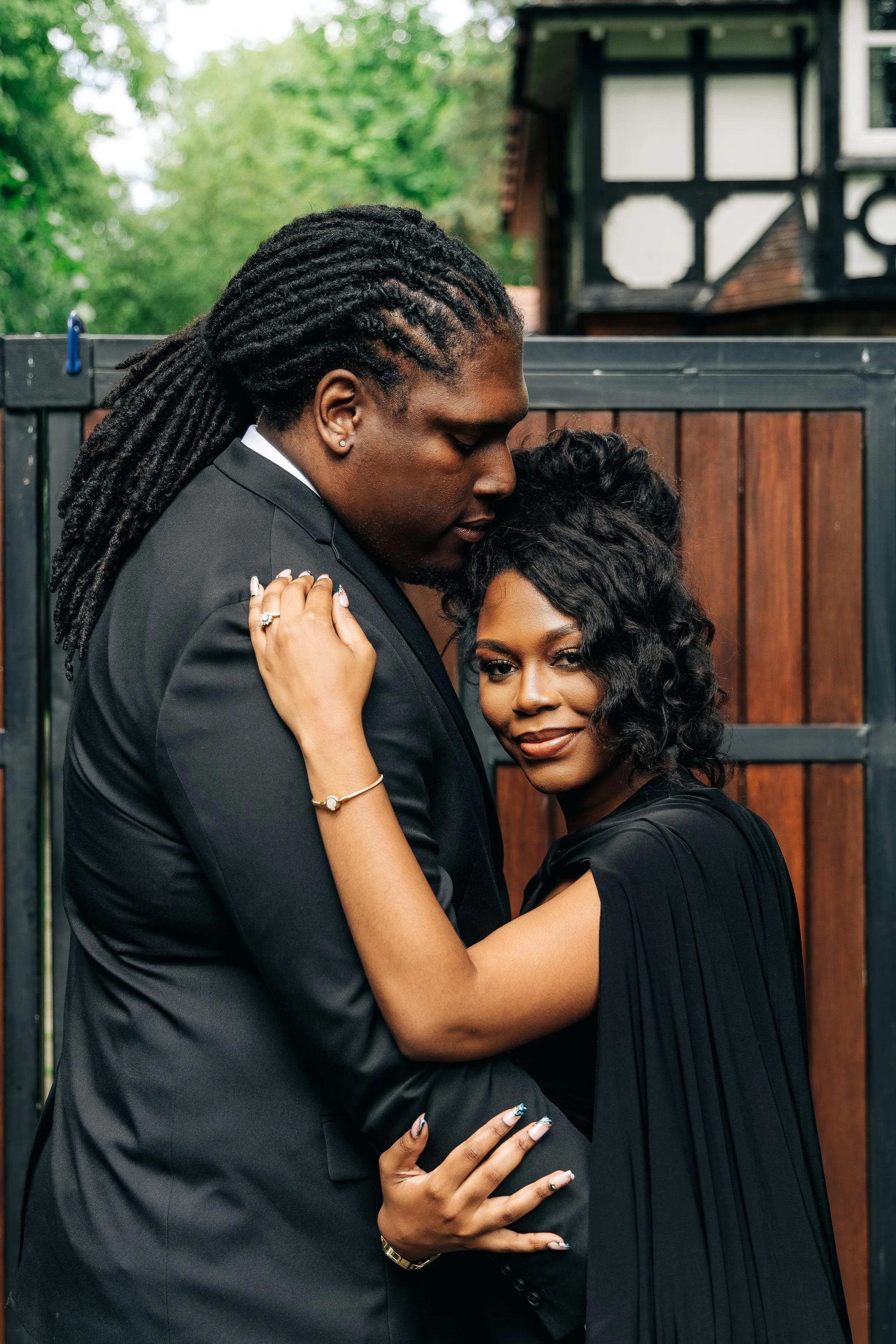 A couple dressed in formal black outfits sharing an intimate moment outdoors in front of a wooden fence with green trees in the background.