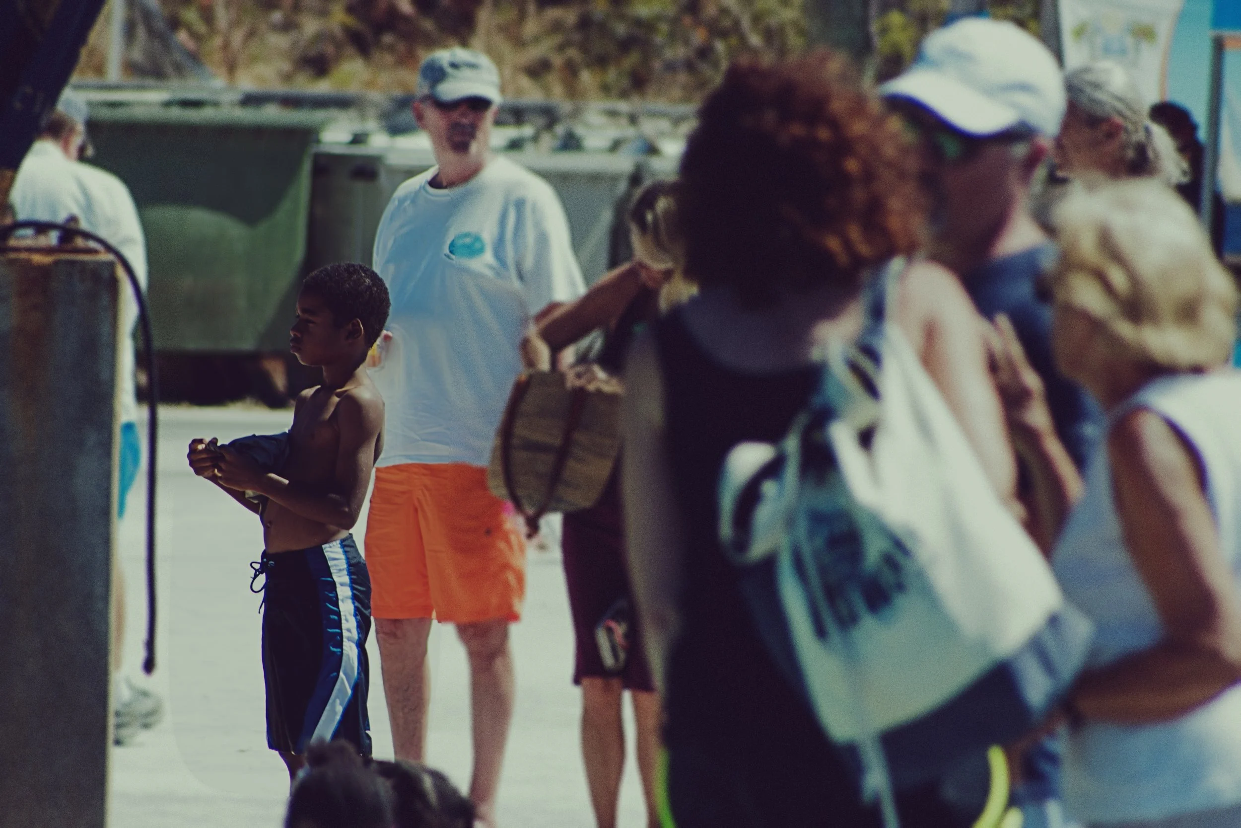 A group of people, including a young boy, standing in line outdoors, some wearing swimwear and caps, with a background of trees and a parking lot.