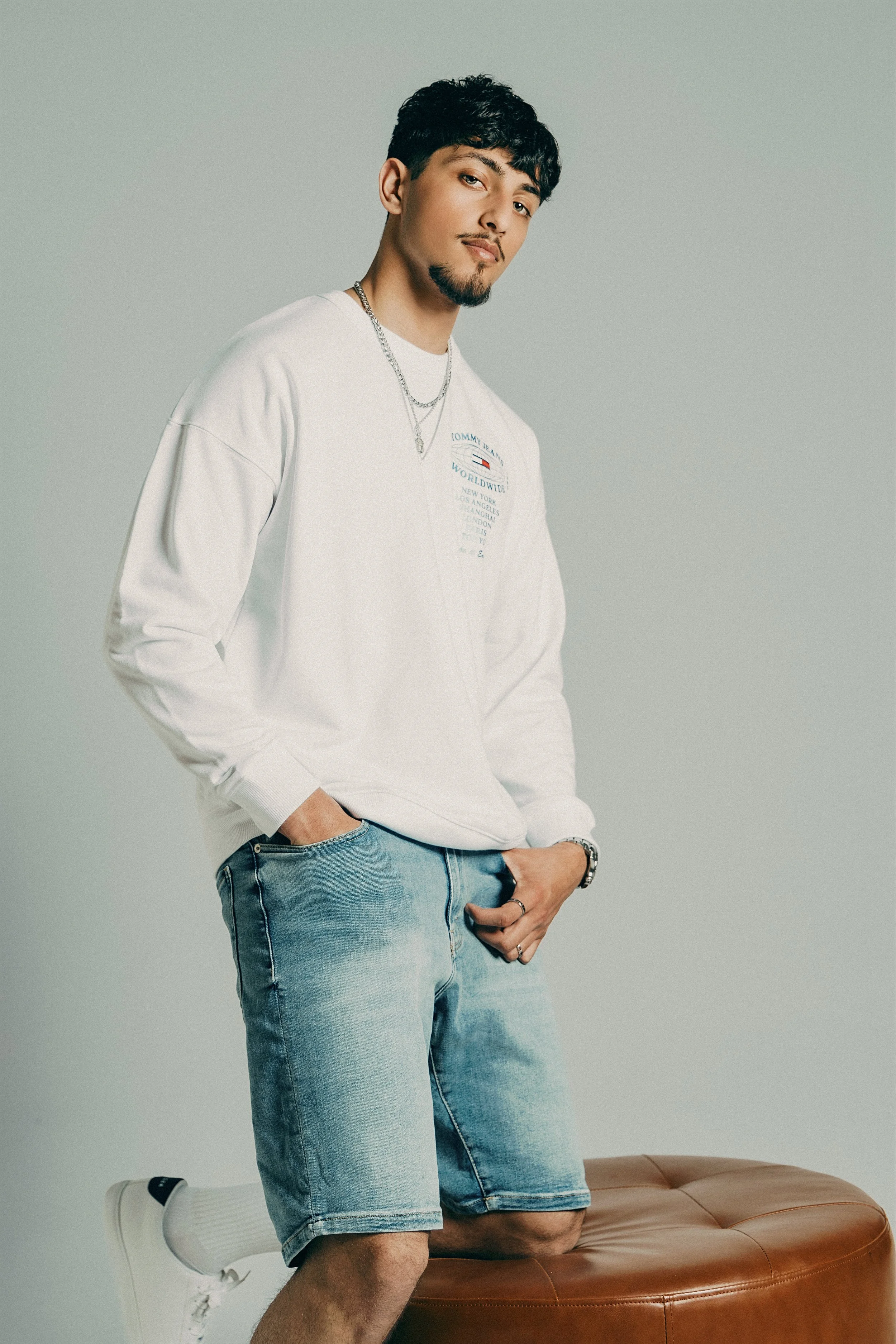 A young man with dark hair, a beard, and mustache, wearing a white Tommy Jeans sweatshirt, light blue denim shorts, white sneakers, and accessories including necklaces, a watch, and rings, posing with one knee on a brown leather ottoman against a pla