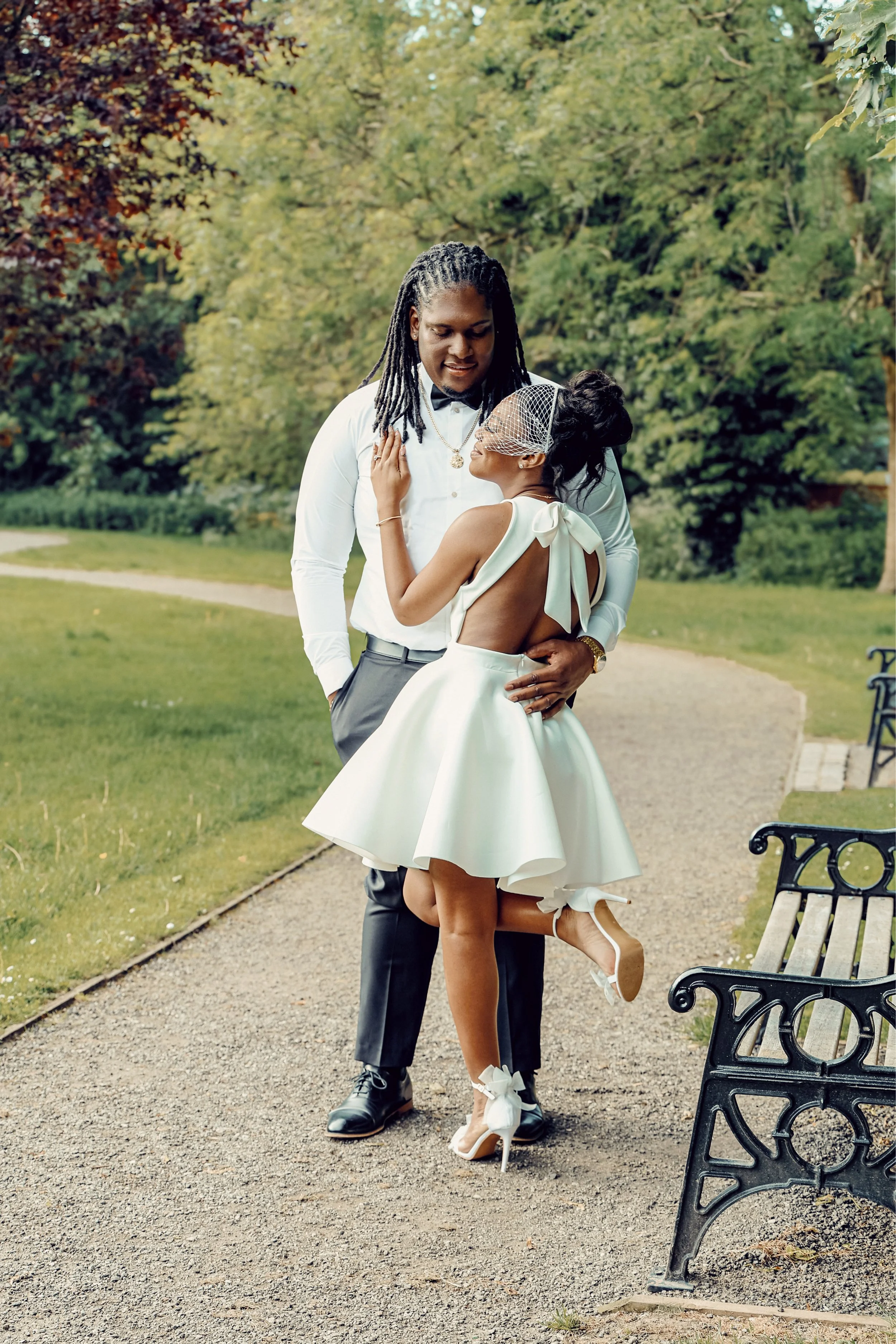 A couple dressed in formal attire, standing on a park pathway surrounded by trees, with the woman lifting one leg and smiling at the man.