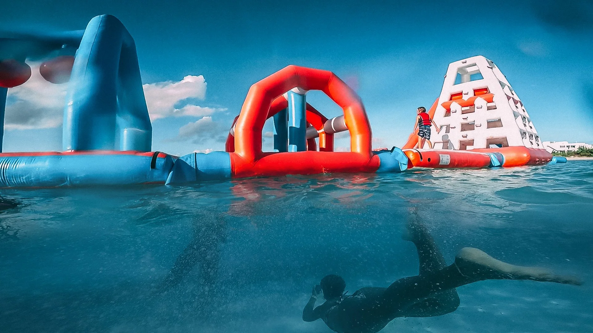Swimmers in a clear blue water pool, with red, blue, and orange floating inflatable obstacle course structures, and a white building in the background.