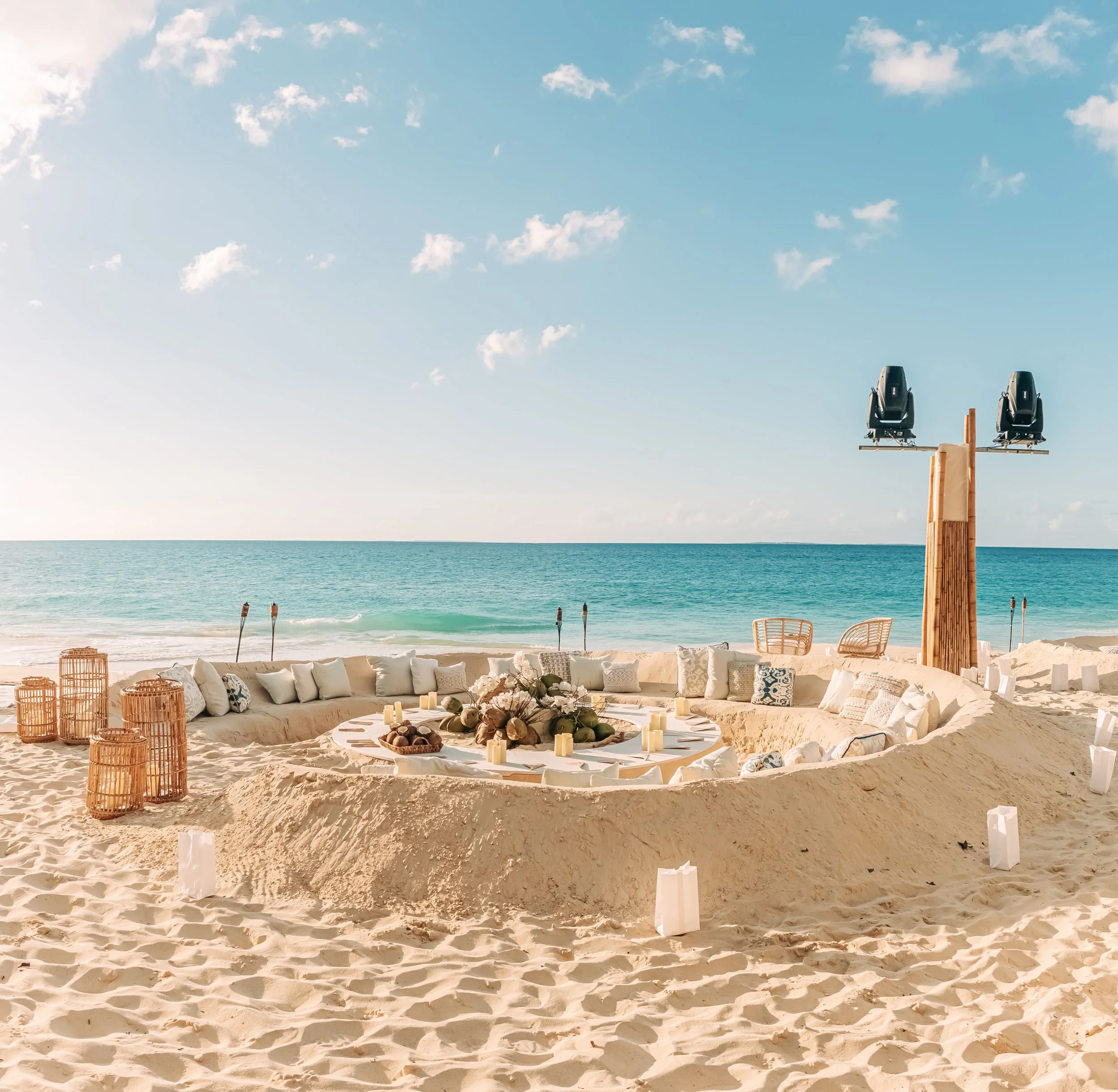 Beach with sandy area, white lounge chairs, and a fire pit with candles, overlooking ocean water under a partly cloudy sky, with two mounted speakers and a bamboo fence.