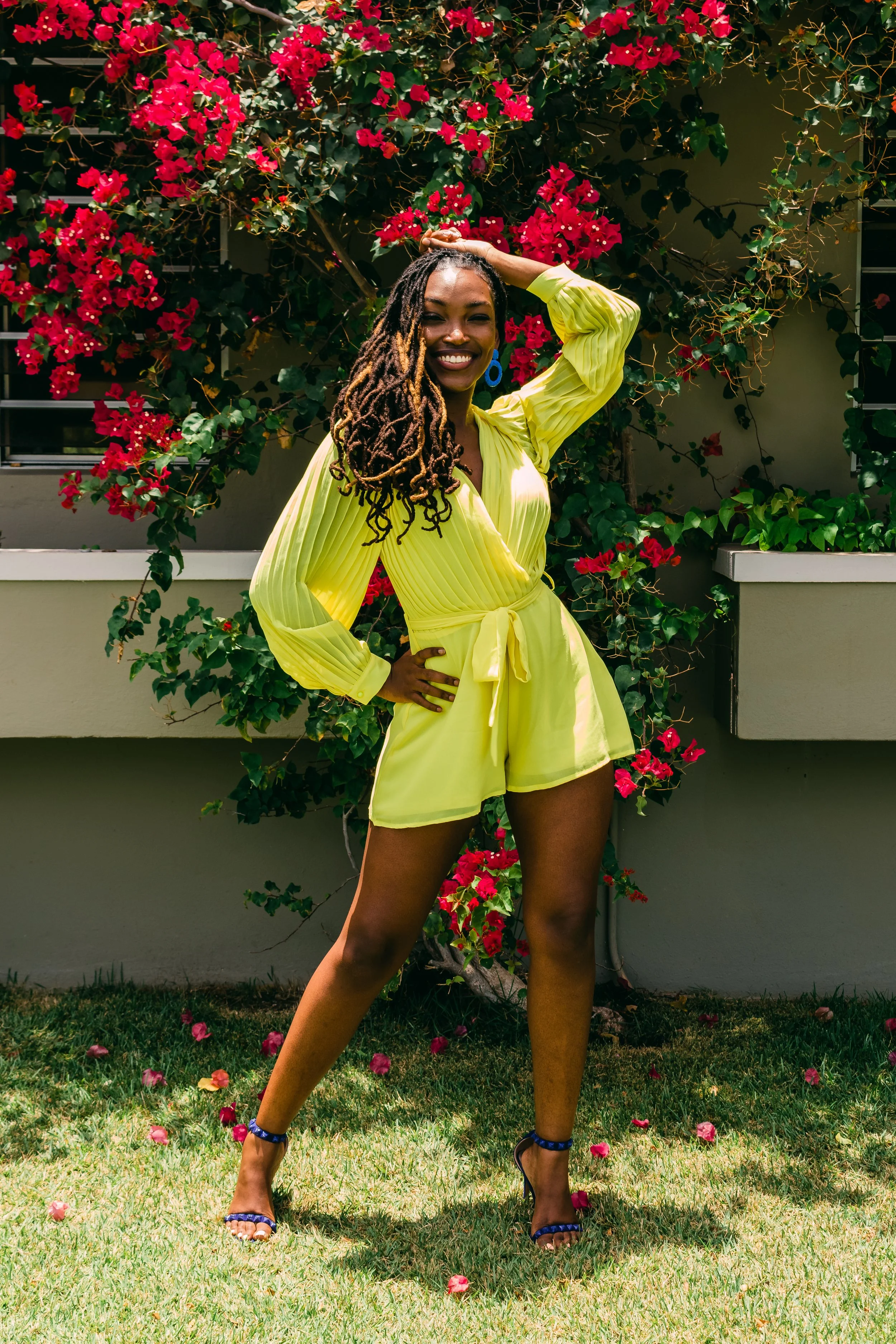 Smiling woman with long dreadlocks posing outdoors in front of a flowering bush with red flowers, wearing a yellow outfit and high heels.