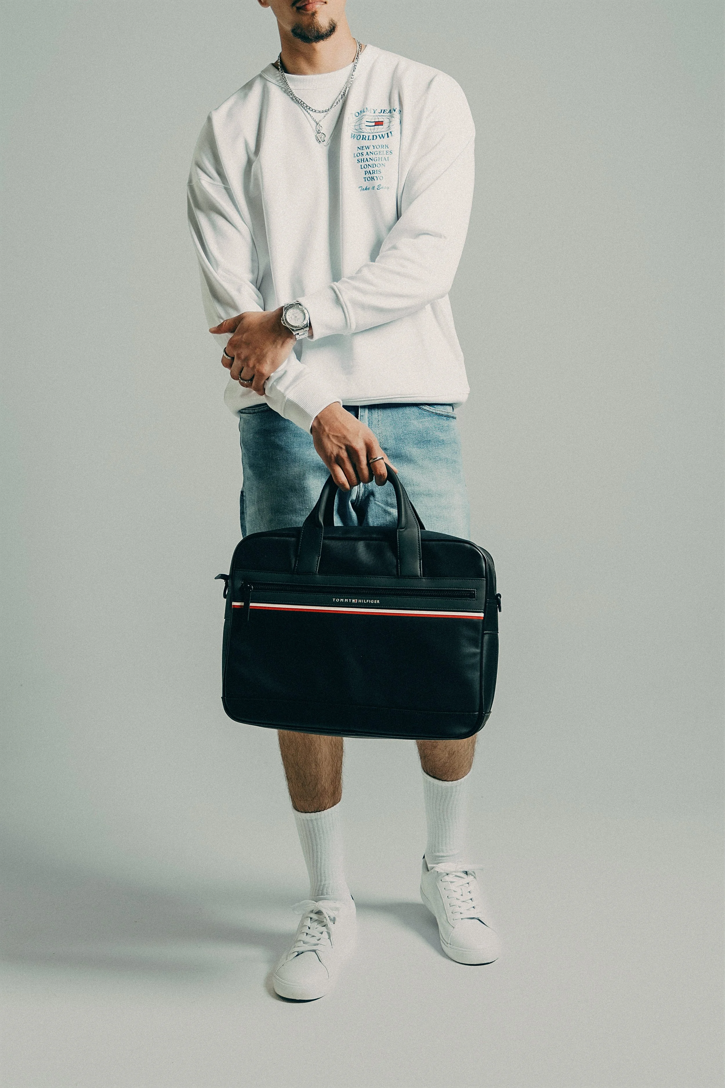 A man in a white sweatshirt, denim shorts, white sneakers, and white socks is holding a black Tommy Hilfiger briefcase, with his left hand crossing his body and resting on his right arm.