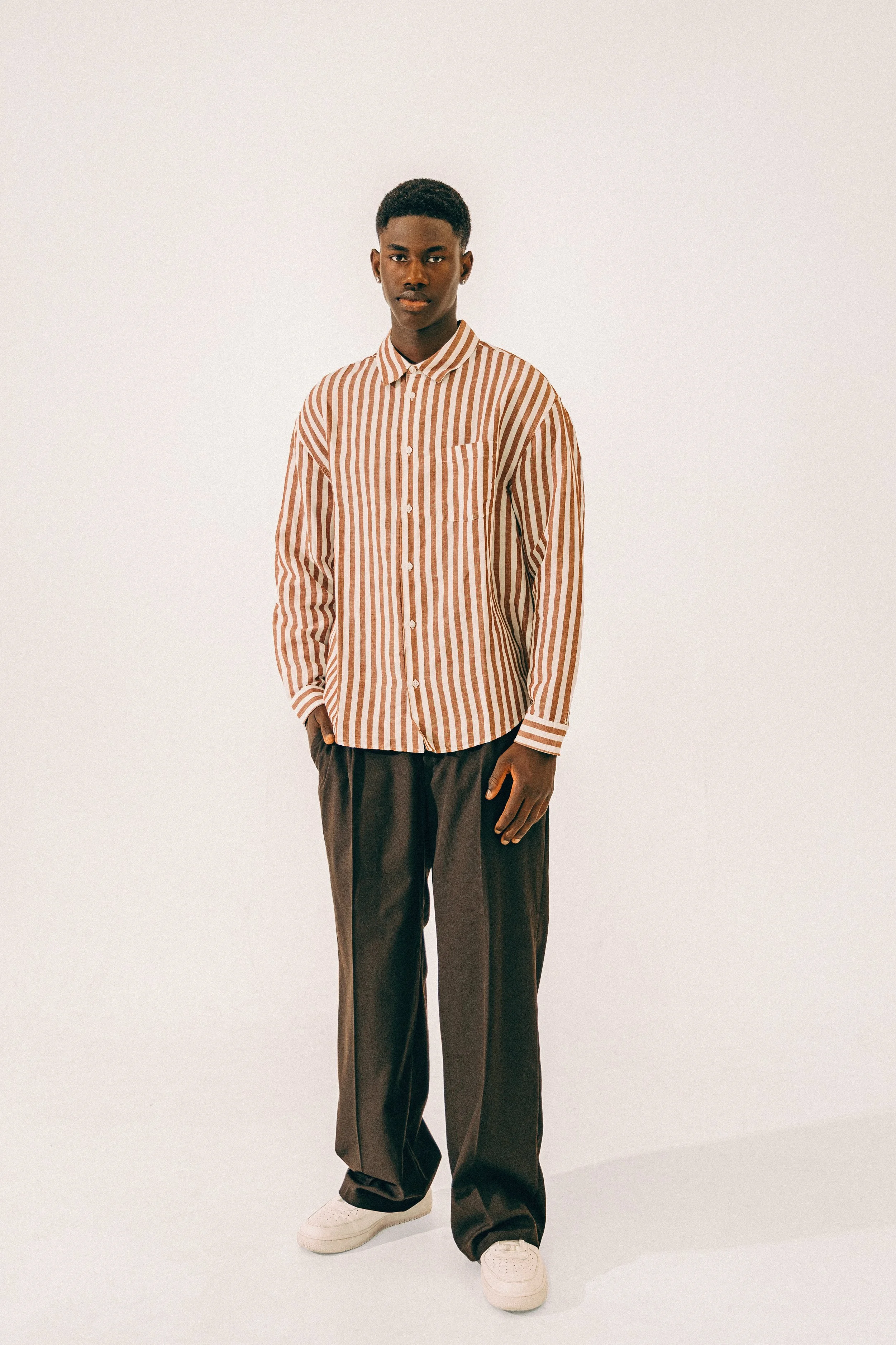 Young man standing against a plain white background, wearing a long-sleeve striped shirt, dark wide-leg trousers, and white sneakers.