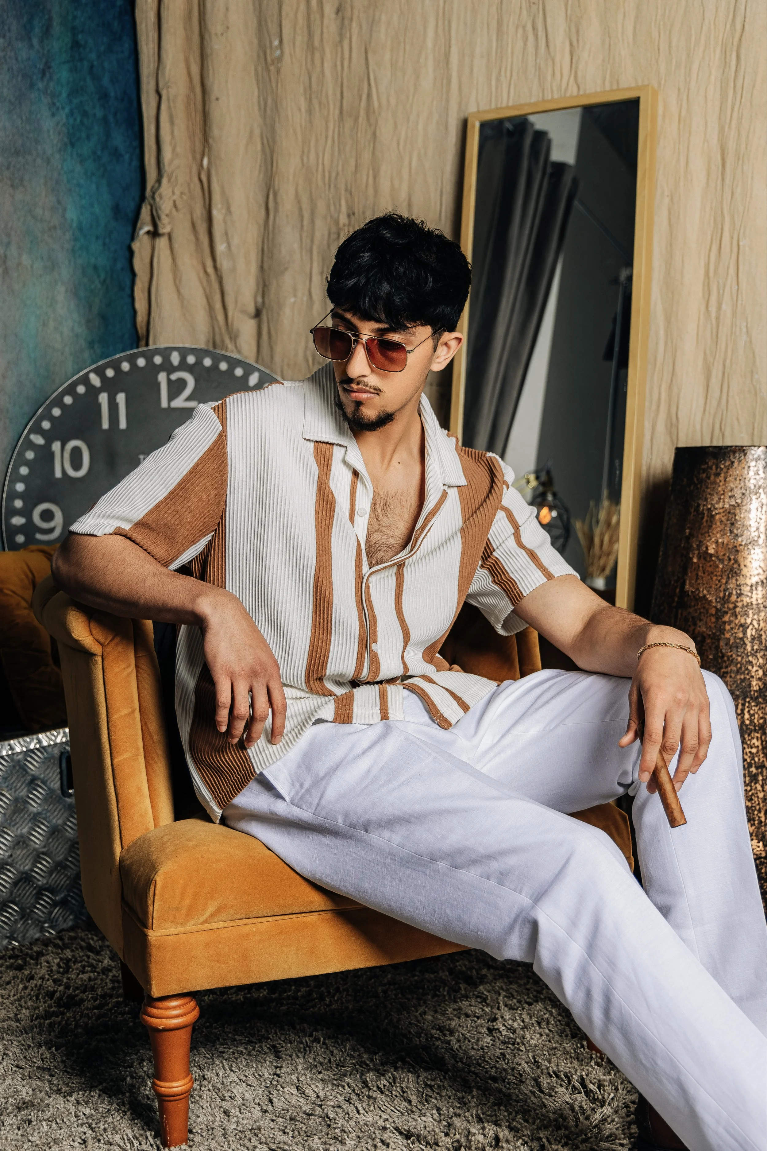 A young man with dark hair, wearing sunglasses, a striped short-sleeve shirt, and white pants, sitting on a mustard-colored vintage armchair, holding a cigar, in a room with a large wall clock, a mirror, and artistic decor.