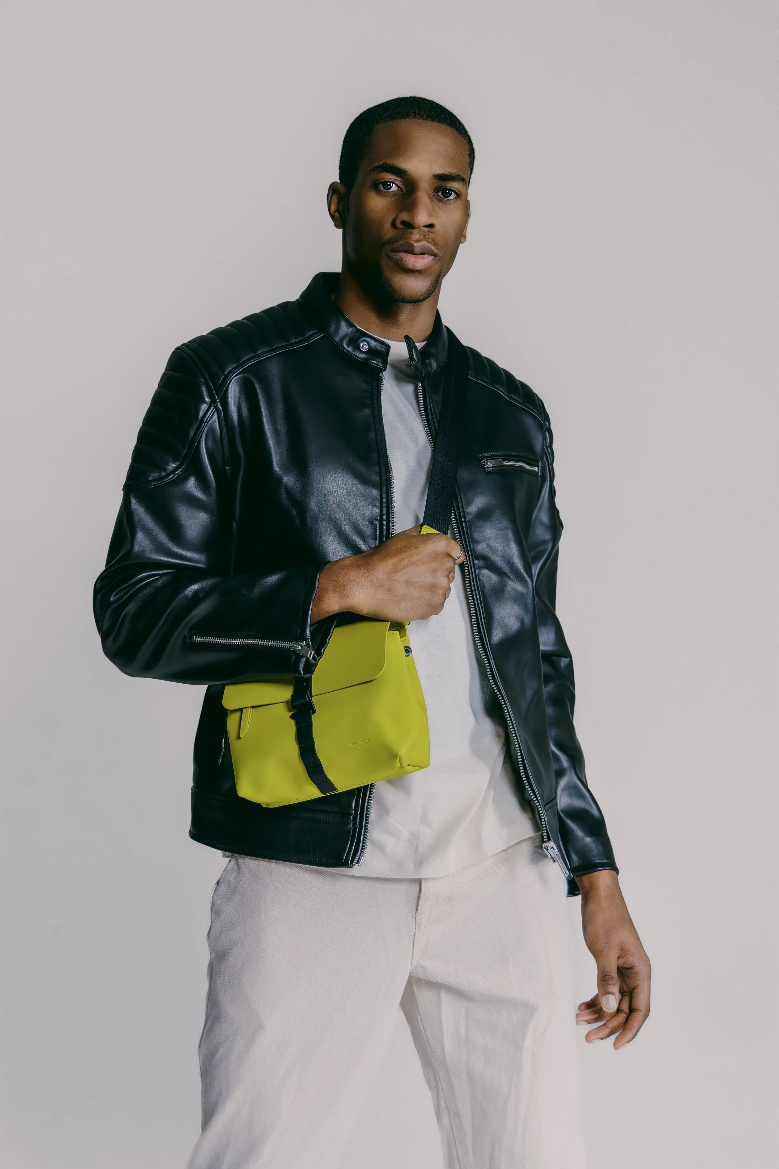 A young man holding a yellow bag, wearing a black leather jacket, gray shirt, and light-colored pants, standing against a plain background.