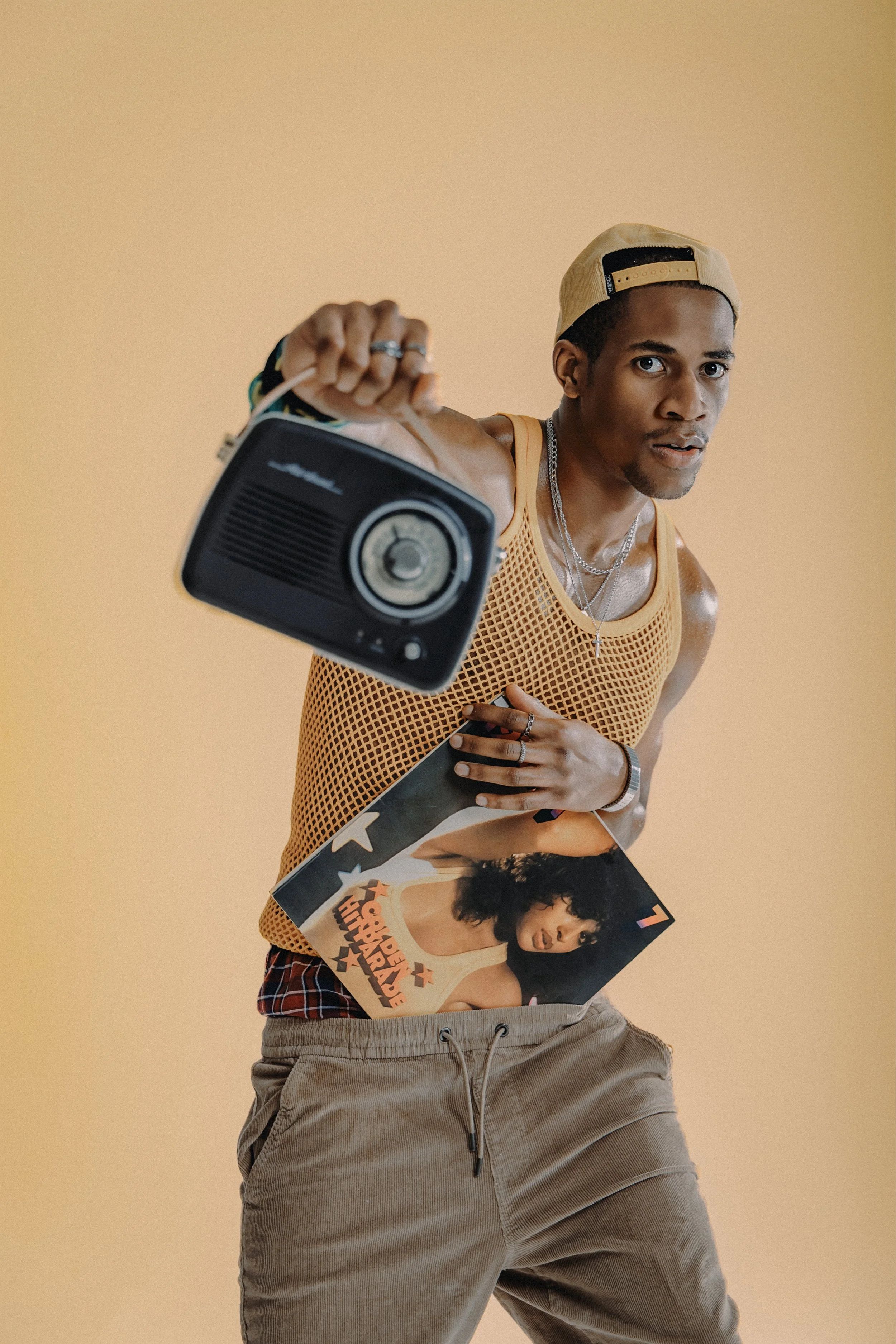 A young man wearing a yellow mesh top, beige cap backwards, and multiple jewelry accessories is holding a vintage radio and a vinyl record with a colorful album cover featuring a woman with curly hair and the text "HOPPER OUTRAGE" against a plain bei