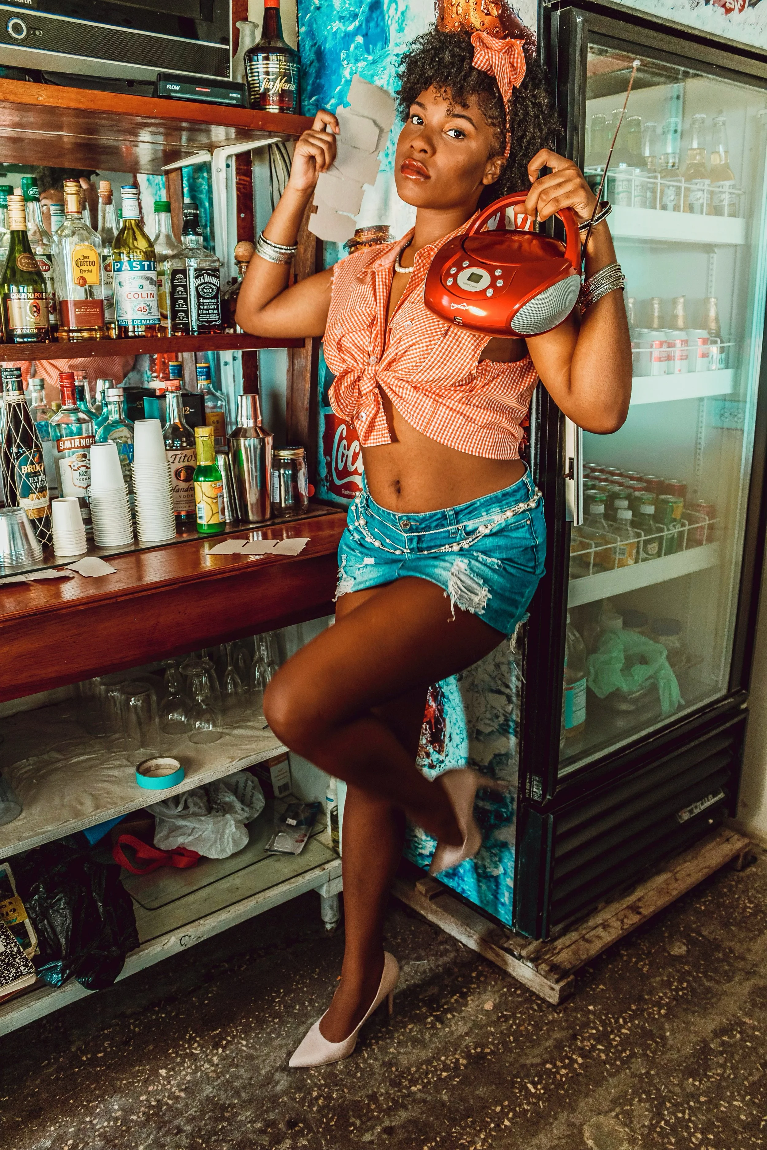 A woman with curly hair in a red band, wearing a knotted red checkered shirt and denim shorts, posing with one leg raised and holding a red boombox in a bar or convenience store setting.