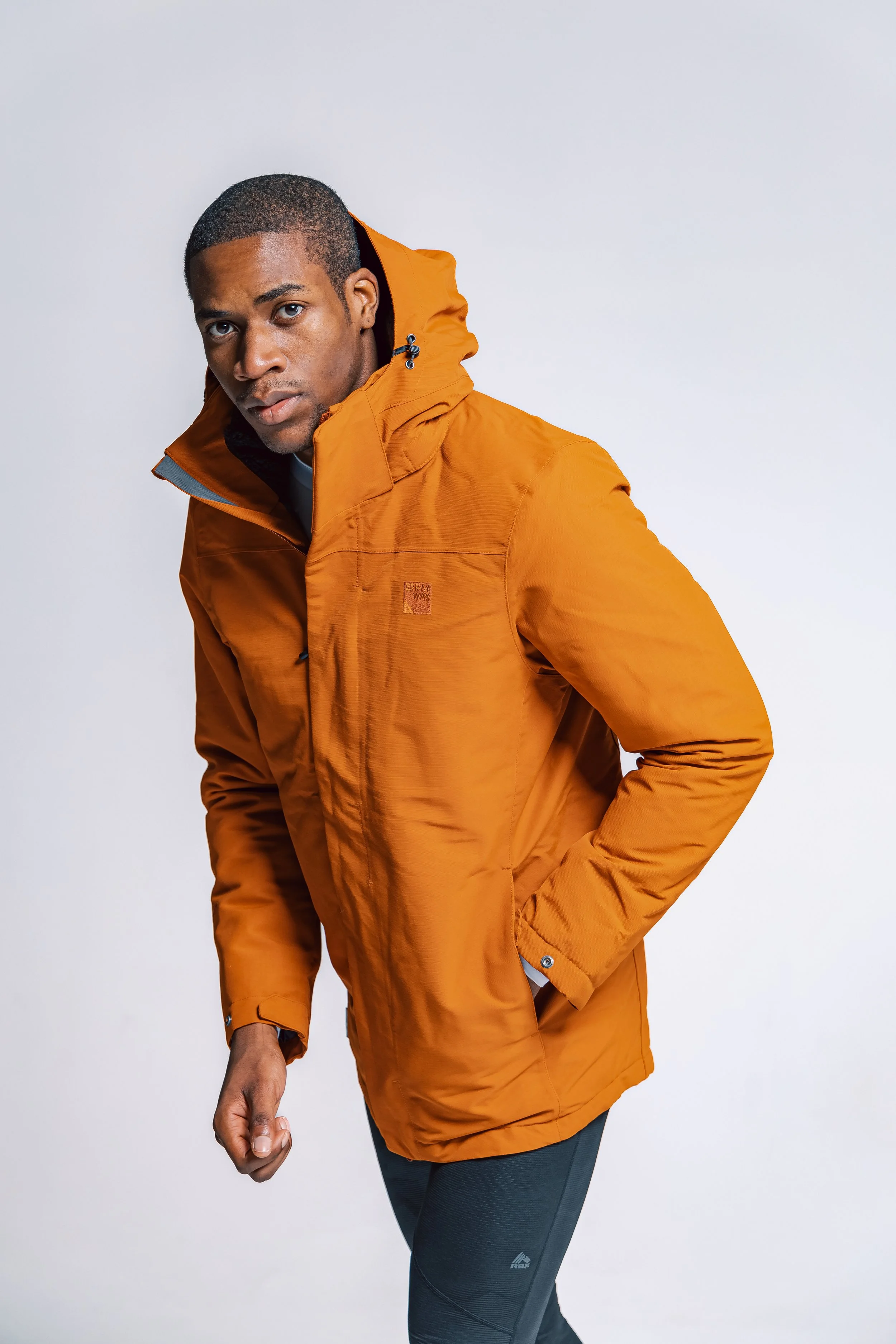Man wearing an orange outdoor jacket and black athletic pants, posing against a plain background.