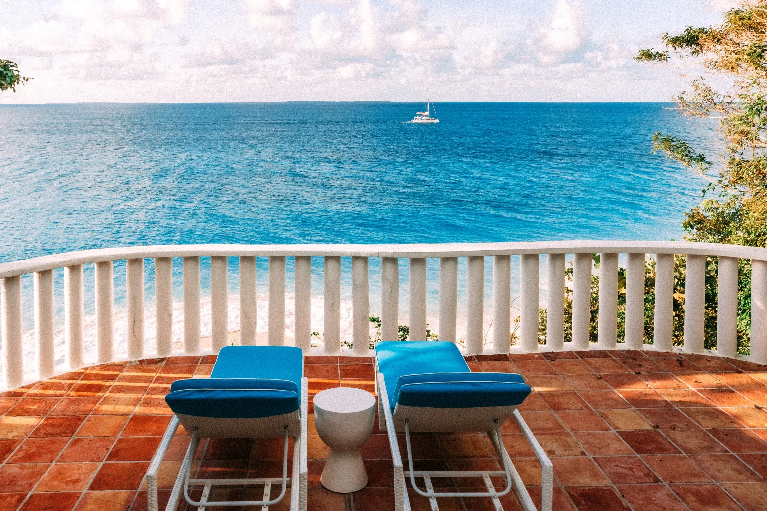 Balcony with blue lounge chairs facing the ocean with a sailboat in the distance, surrounded by trees and a white railing