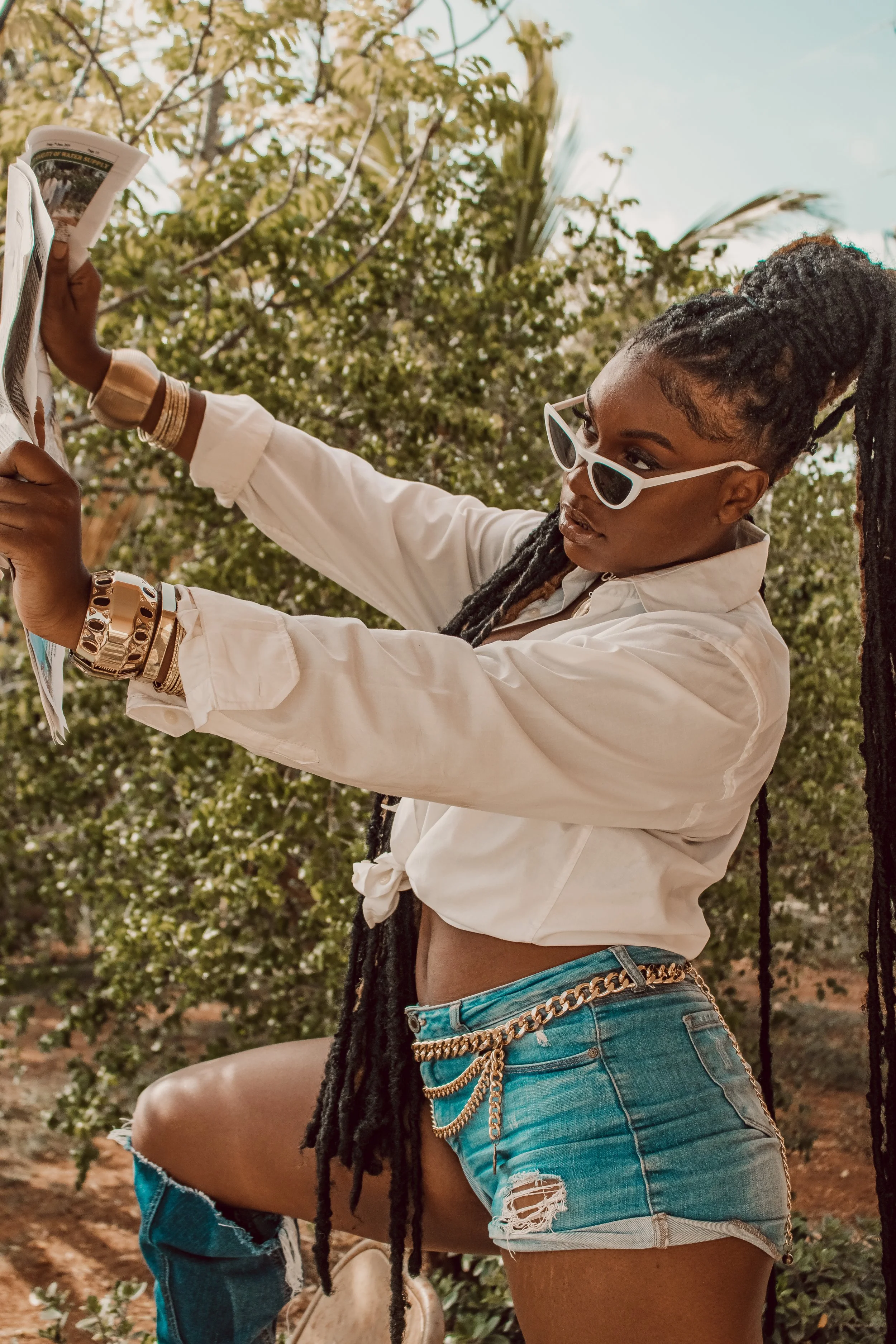 A woman with dreadlocks wearing white sunglasses, a white cropped shirt, and ripped denim shorts with chains, standing outdoors among greenery, holding and looking at a magazine.