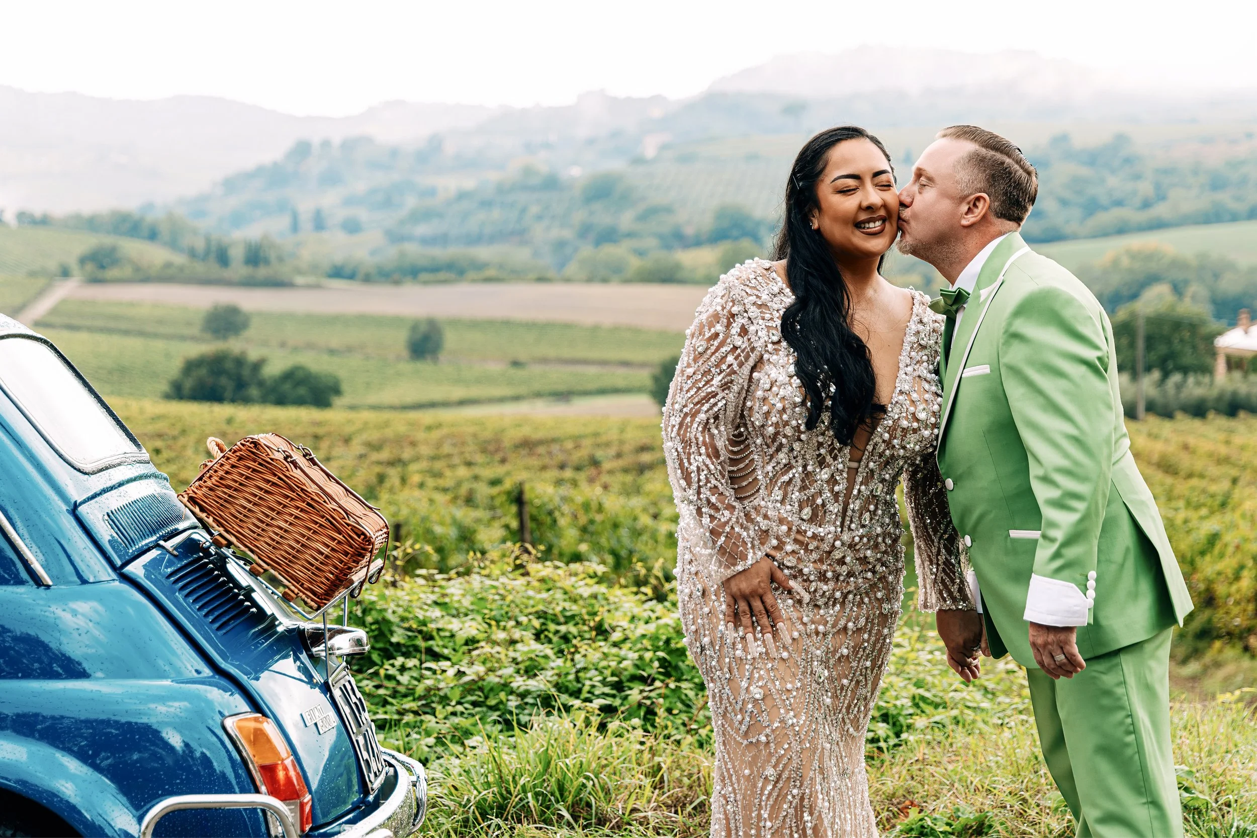 A couple dressed in elegant wedding attire with a woman in a beaded gown and a man in a bright green suit, standing in a vineyard field during daytime. The man is kissing the woman on the cheek, and there is a vintage blue car with a wicker picnic ba