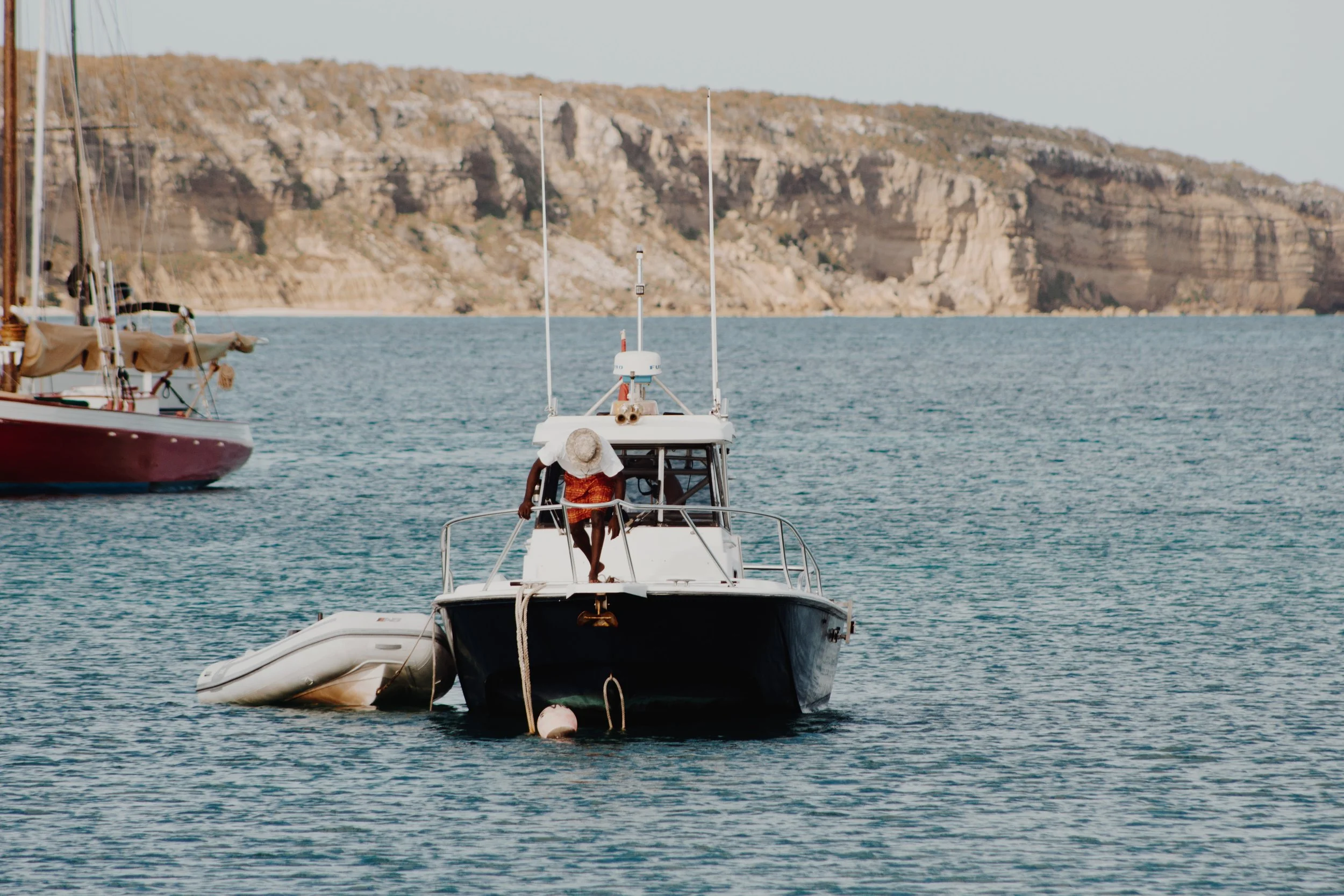 A person wearing a hat sitting on the back of a boat in a harbor, with sailboats and rocky cliffs in the background.