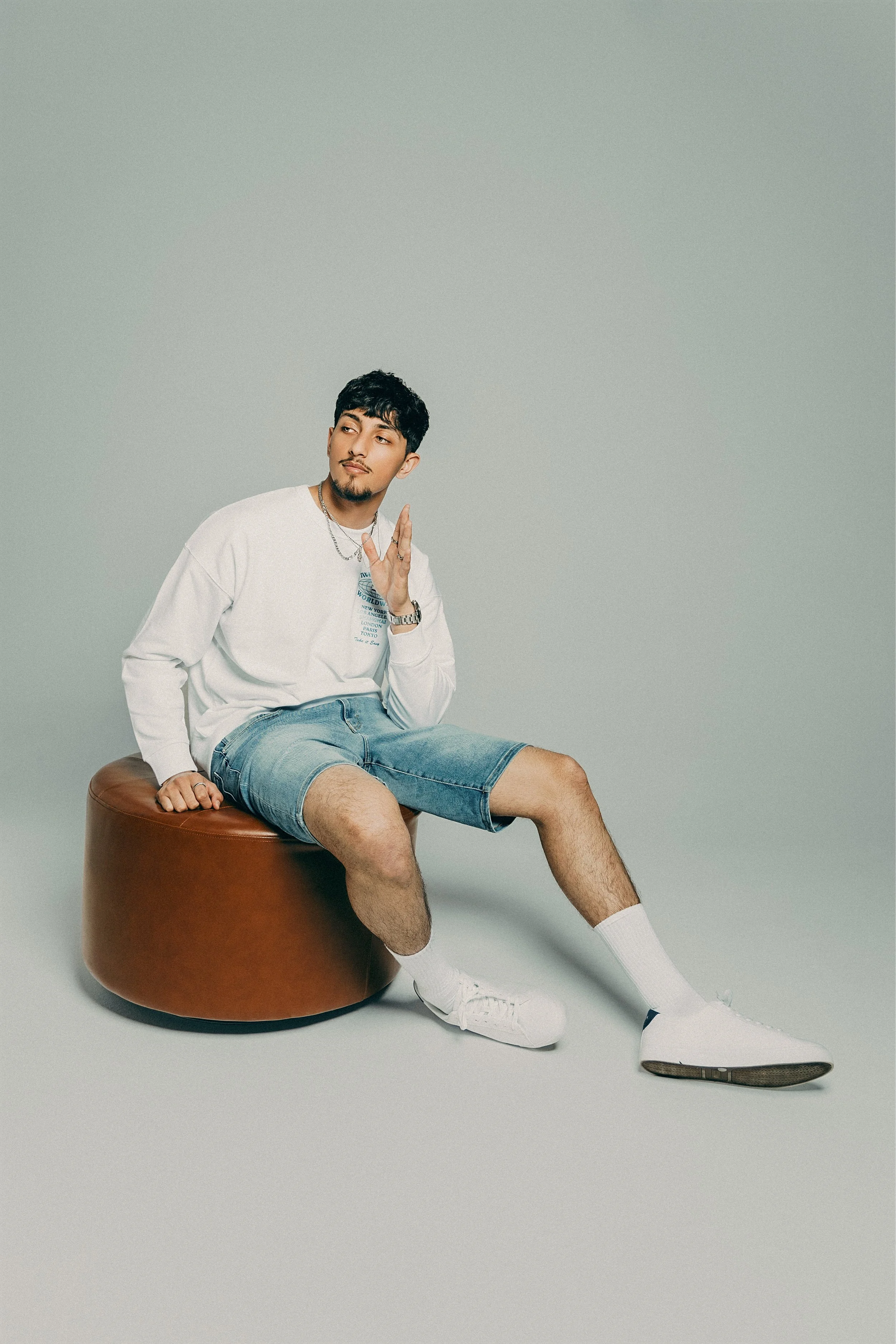 A young man with black hair, facial hair, and light skin sits on a round, brown ottoman against a plain, light grey background. He wears a white long-sleeve shirt, denim shorts, white sneakers, and white socks. He looks to the side with his right han