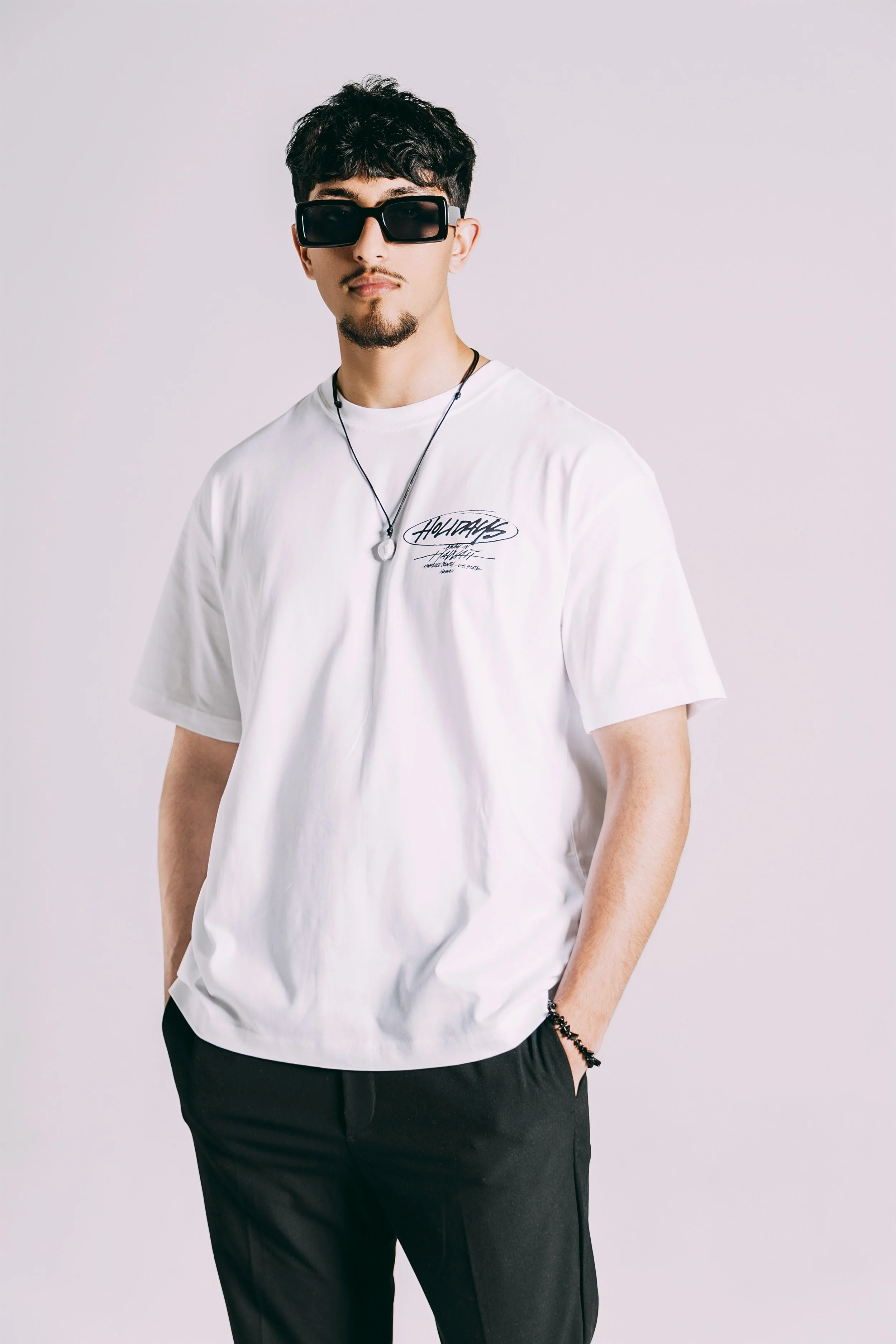 A young man with dark hair, wearing sunglasses, a white graphic T-shirt, black pants, and accessories, stands against a plain white background.