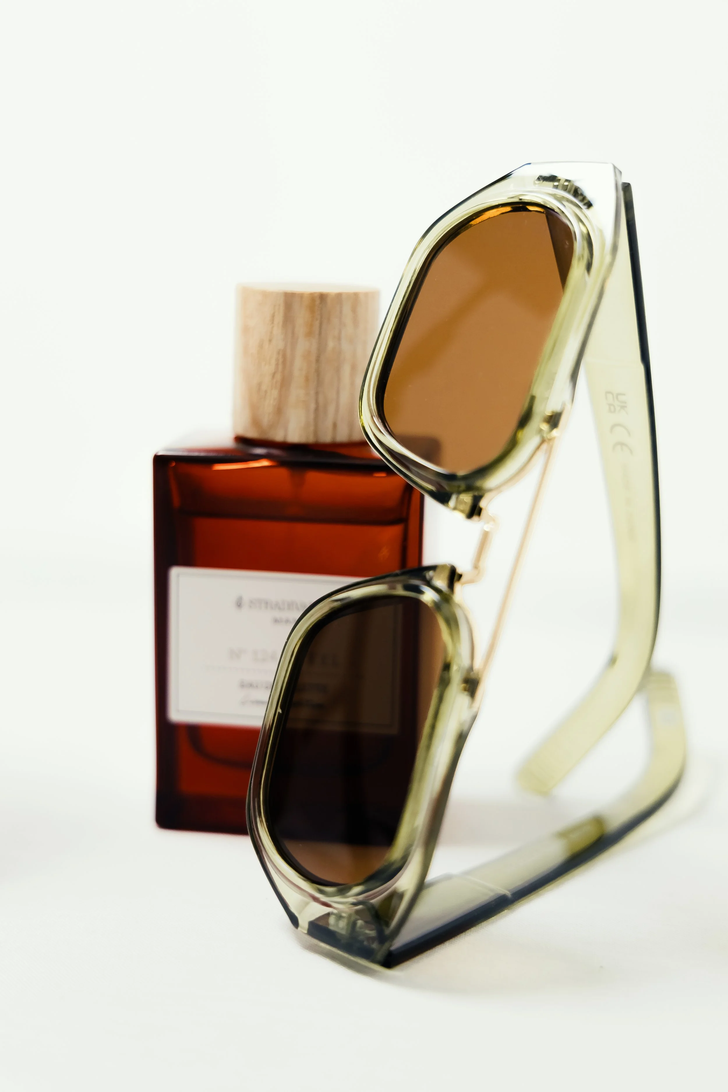 Sunglasses with tinted lenses, a brown perfume bottle with a white label, and a round wooden cap on the bottle, set against a plain white background.