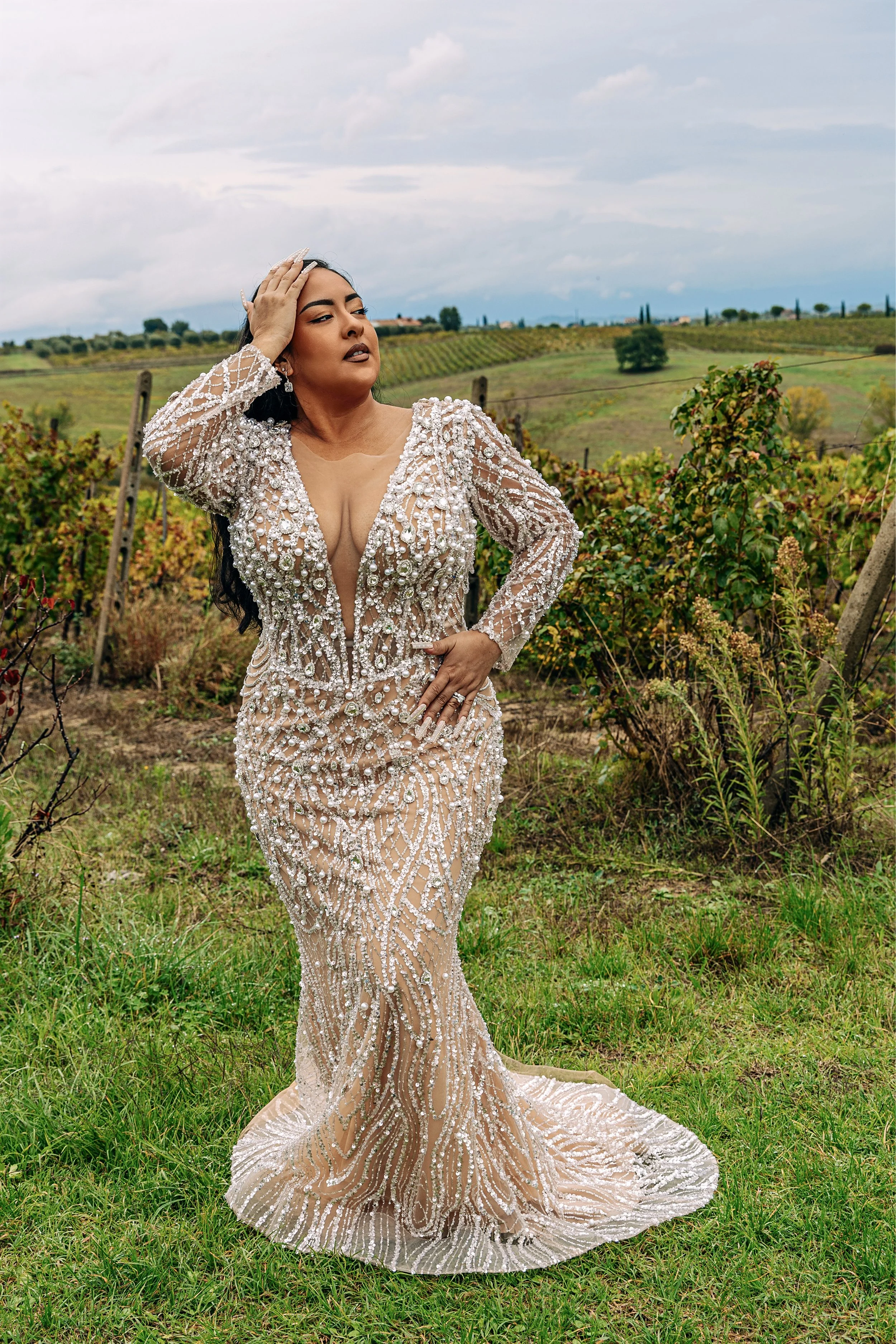 A woman in an elaborate, beaded gown stands in a vineyard with rolling hills and cloudy sky in the background.