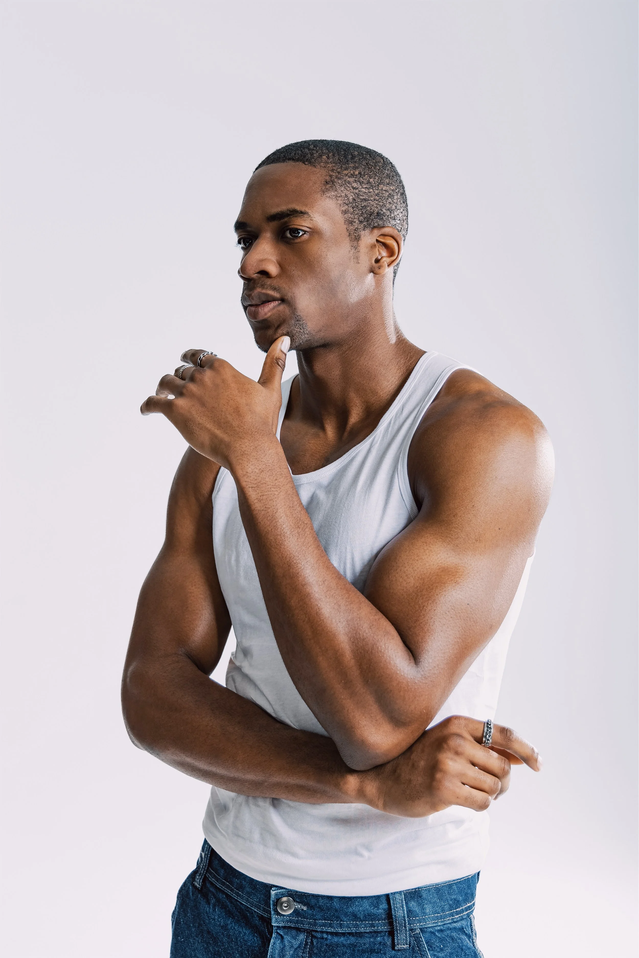 A young man with dark skin and short hair, wearing a sleeveless white shirt and blue jeans, standing against a plain white background. He is touching his chin with his right hand in a thoughtful pose.