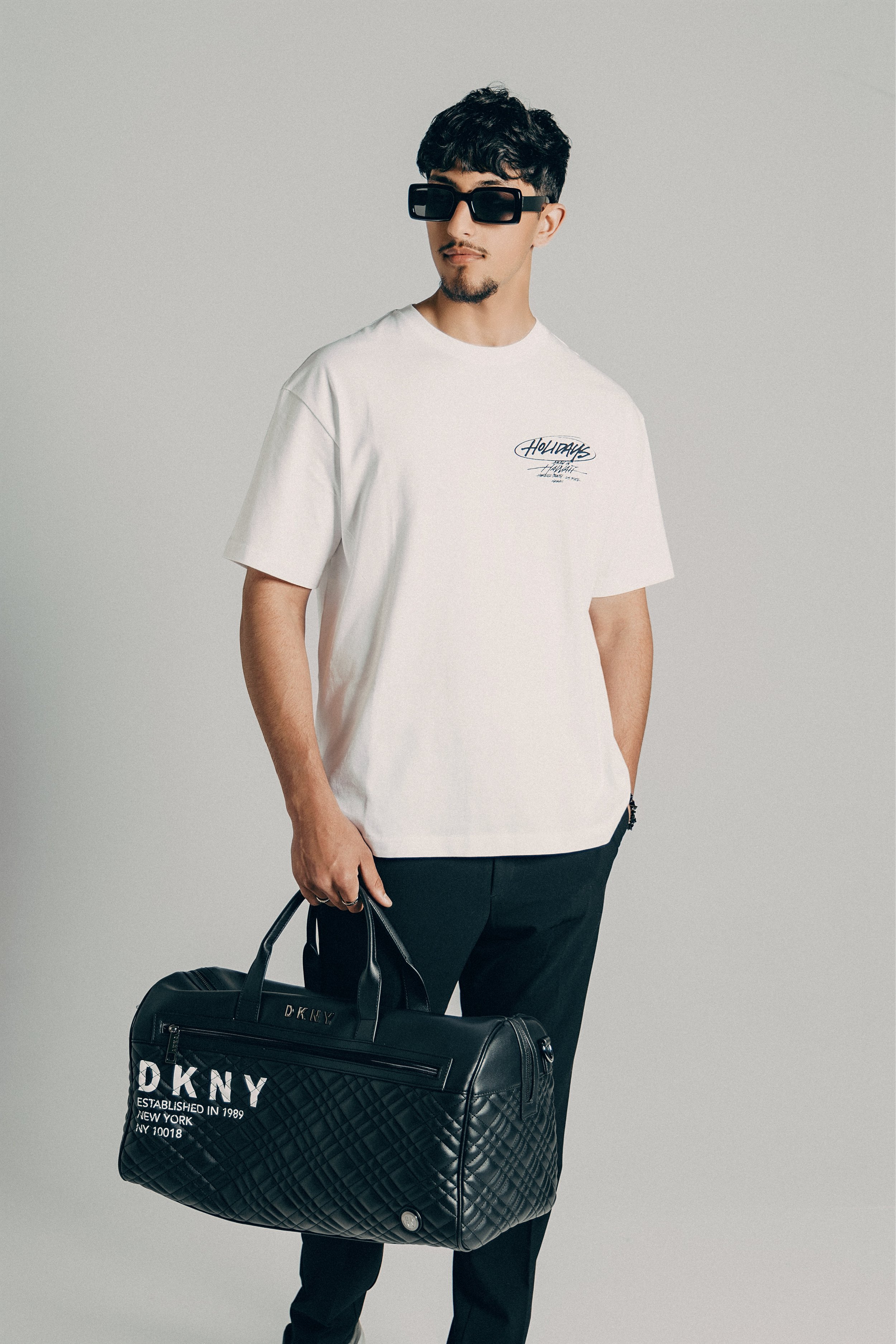 Young man wearing black sunglasses, a white T-shirt with a graphic print, black pants, holding a black DKNY duffel bag, standing against a plain light gray background.