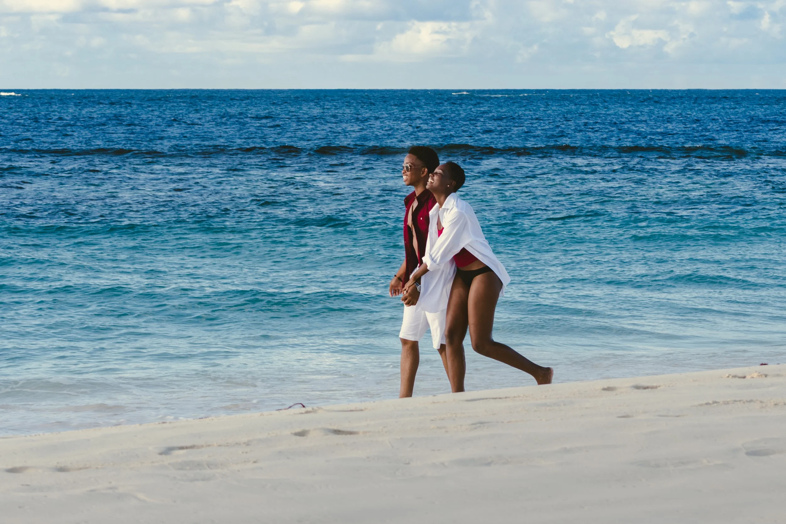 A smiling couple walking along the beach near the ocean, with waves and blue sky in the background.